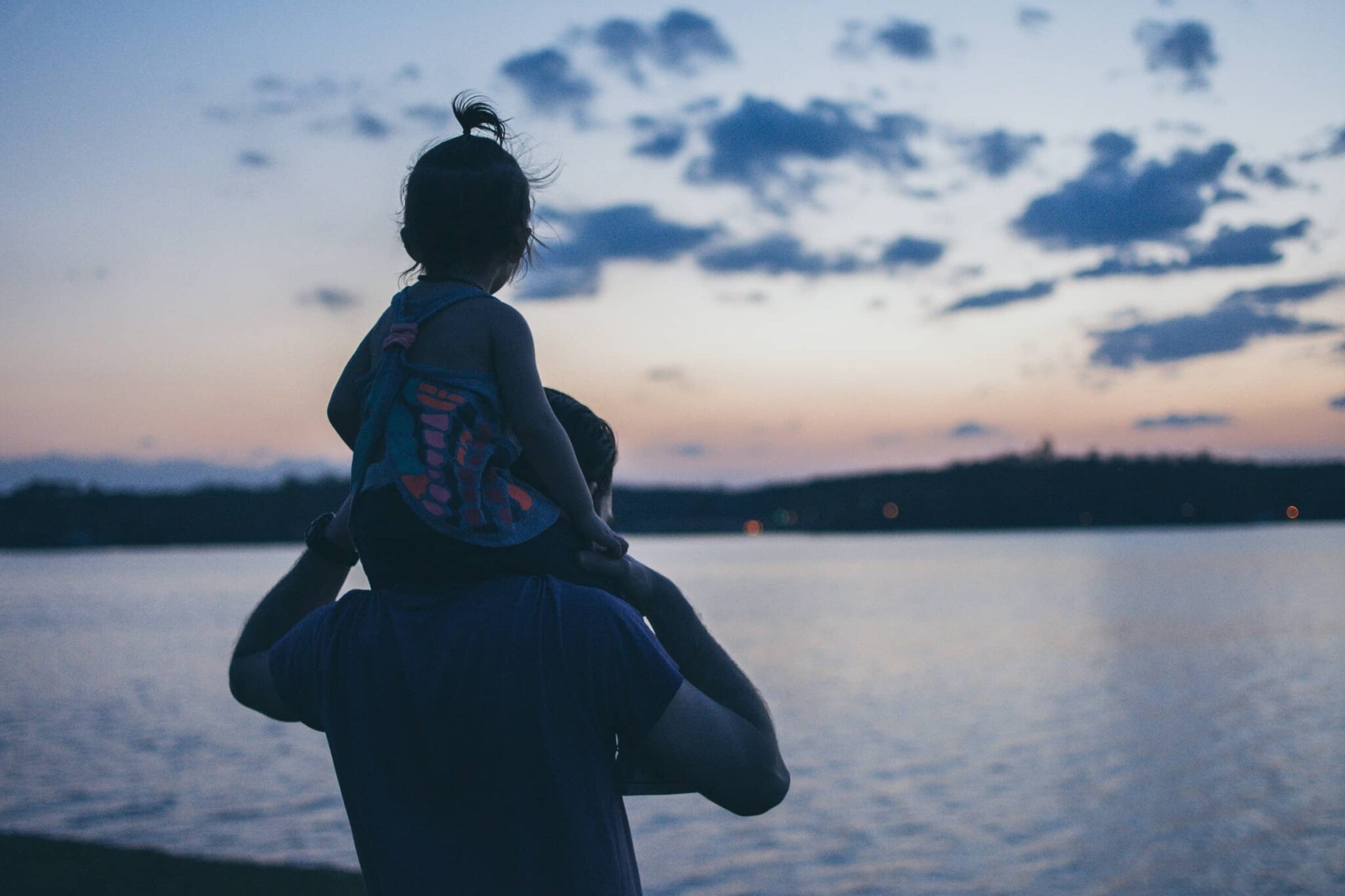 An image of a man with a child on his shoulder in silhouette from behind on a sunset beach.