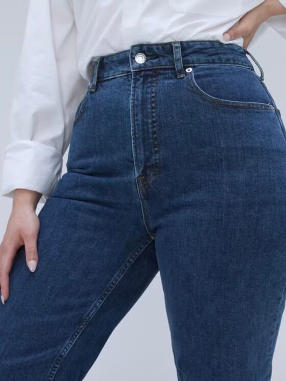 A person wearing a white shirt and high-waisted blue jeans stands with one hand on their hip.