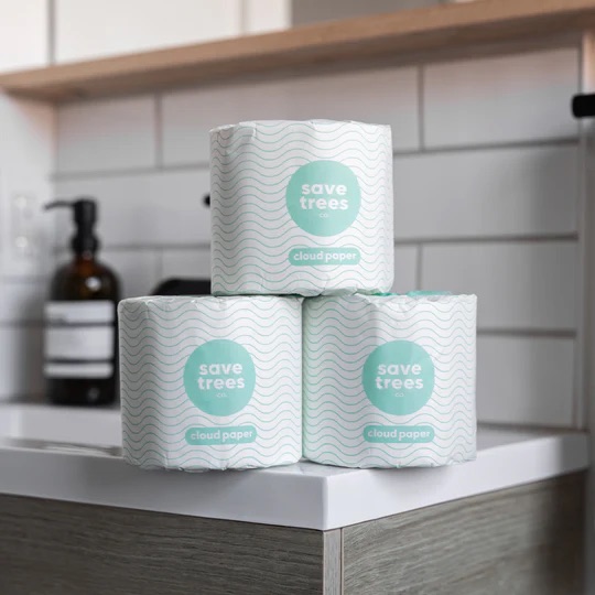 Three rolls of Save Trees brand toilet paper are stacked on a bathroom counter with tiled walls and a soap dispenser in the background.