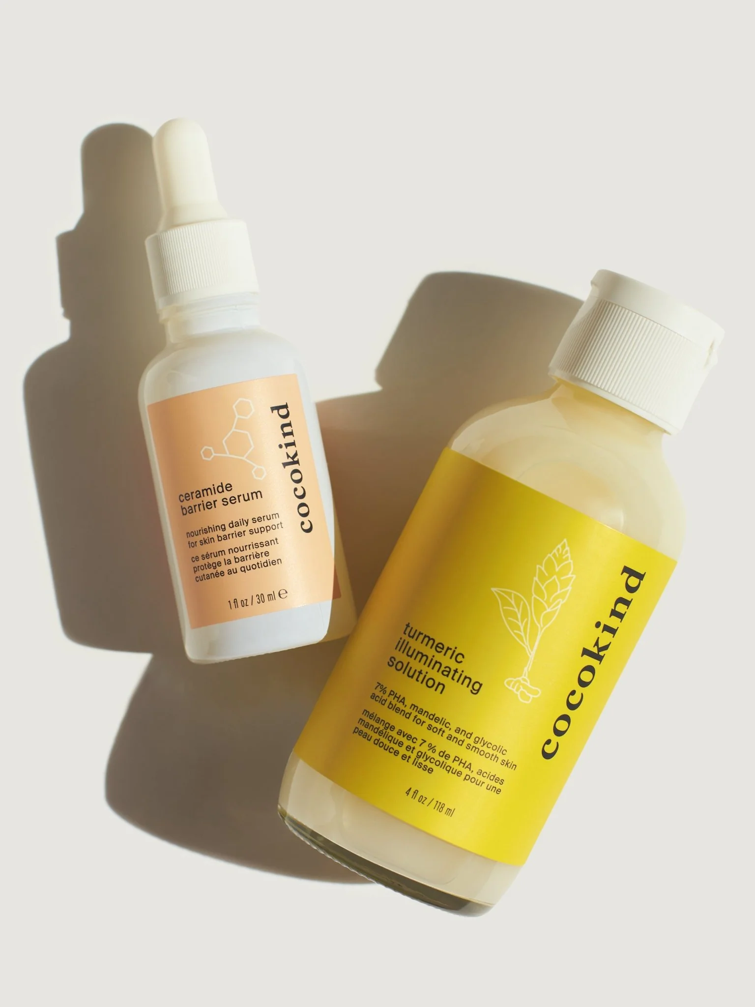 Two skincare bottles on a light background: a dropper bottle labeled "ceramide barrier serum" and a larger bottle labeled "turmeric illuminating solution" by Cocokind.