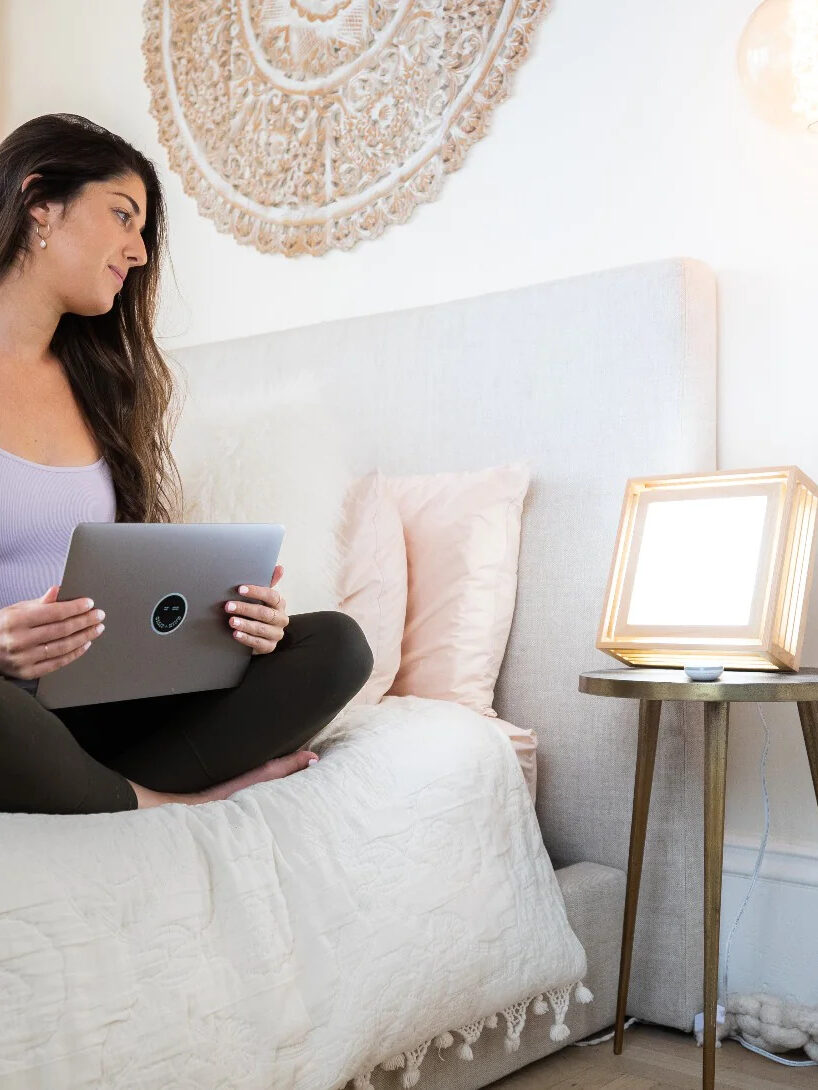 A model seating on a bed, looking at their Sunrise Sensations Lamp. 