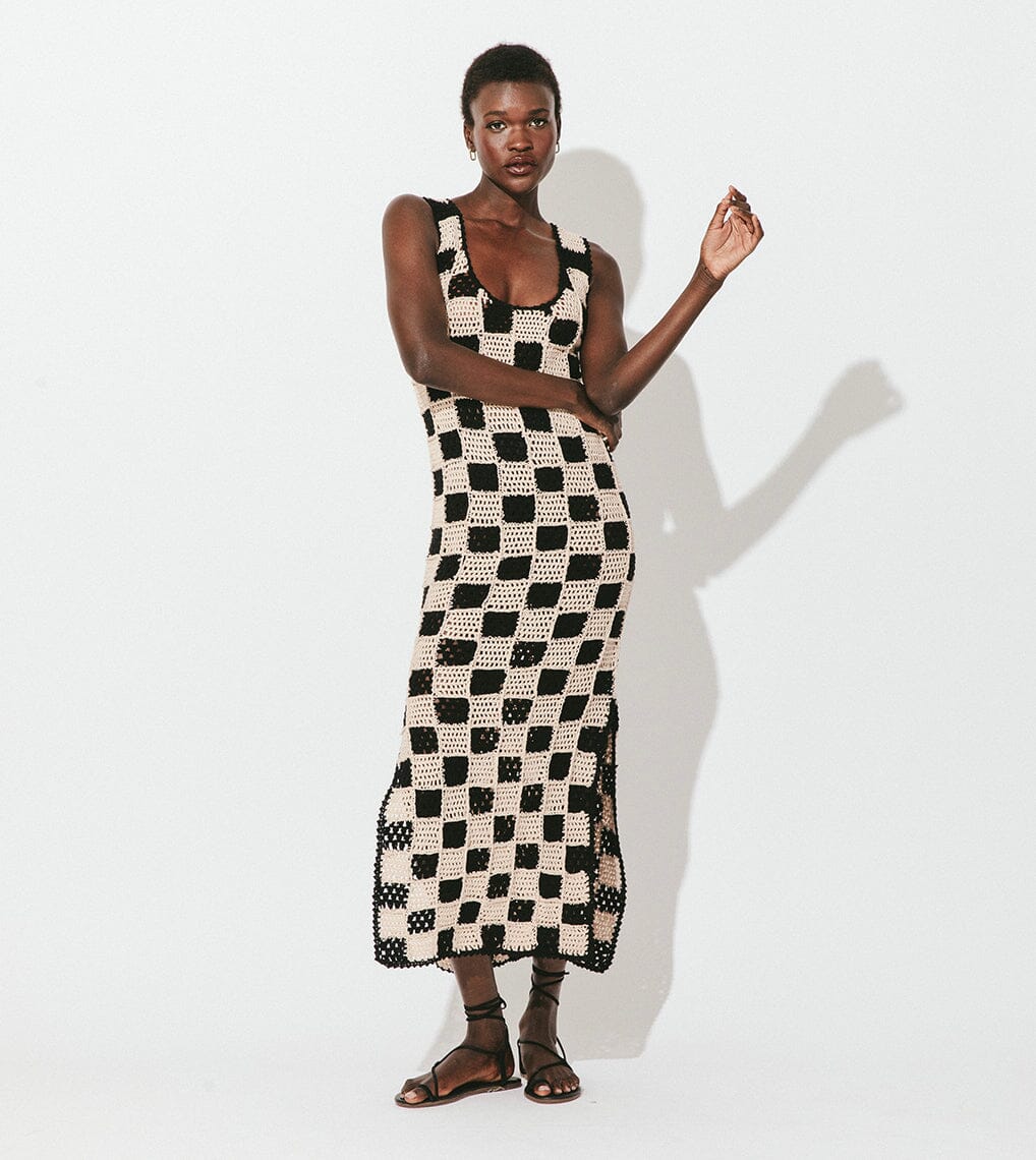 Person wearing a sleeveless black and beige checkerboard-patterned knit dress and black sandals, standing against a plain white background.