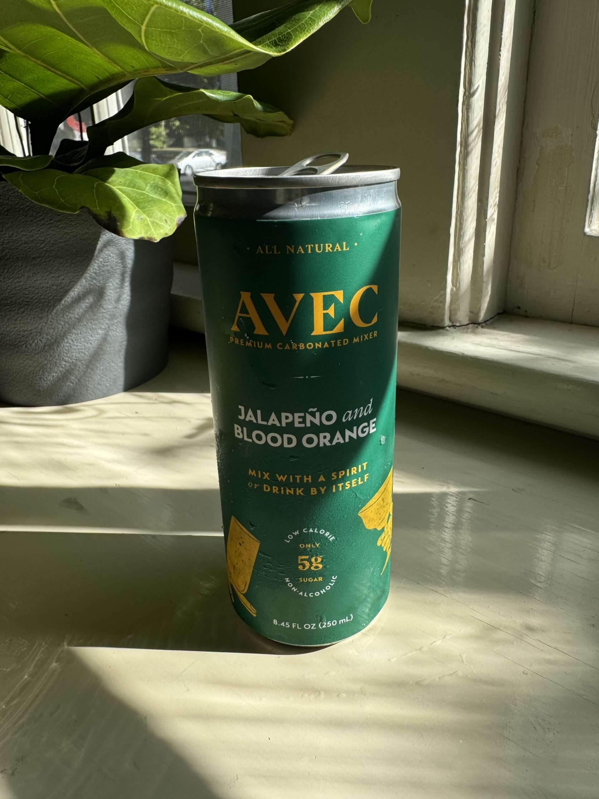 A can of AVEC Jalapeño and Blood Orange premium carbonated mixer sits on a windowsill in sunlight. The green can highlights its all-natural ingredients and suggests mixing with a spirit or drinking by itself.