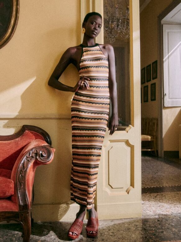 Person standing in a hallway, wearing a sleeveless striped maxi dress and red woven sandals, with one hand resting on a red upholstered chair.