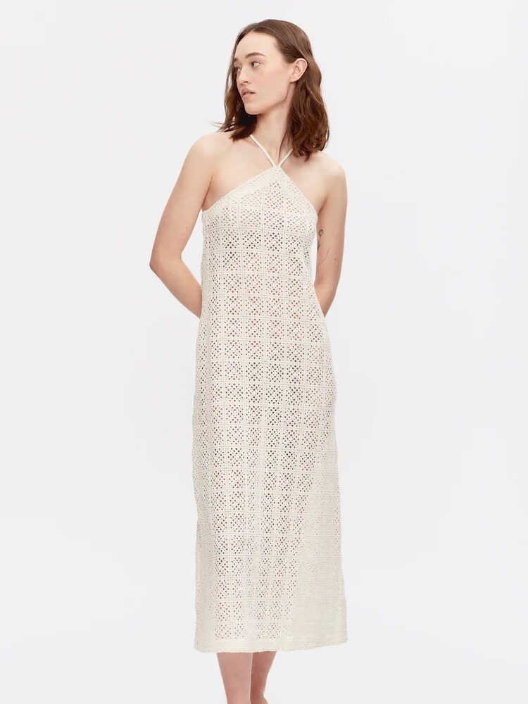 A person is wearing a sleeveless, off-white crochet halter dress, standing with hands at sides against a plain white background.