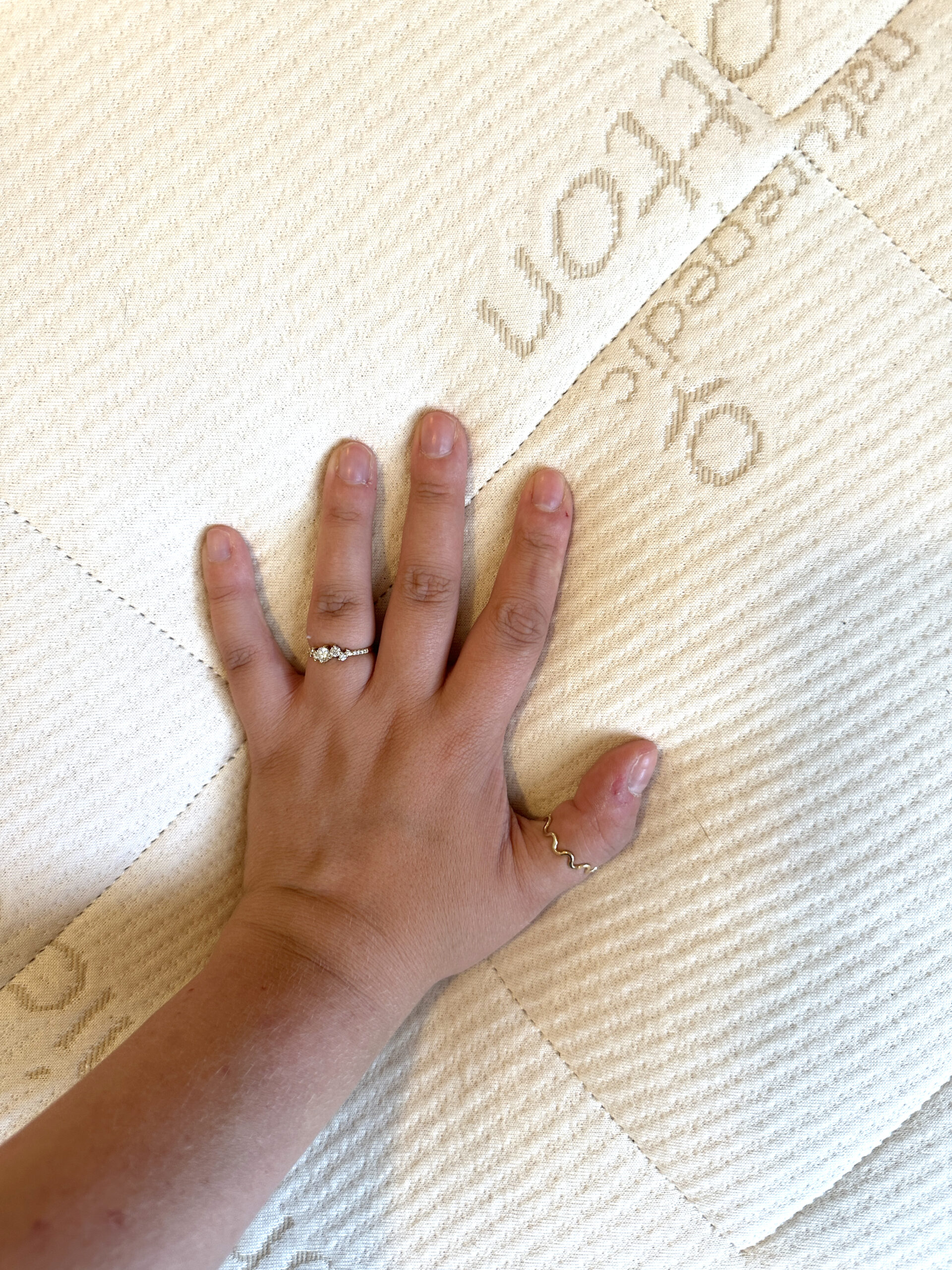 A hand with two rings presses down on a cream-colored, textured mattress with stitched patterns and partial text visible.