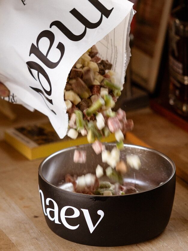 A person pours dry pet food from a large white bag into a black bowl labeled "Maev" on a wooden countertop.