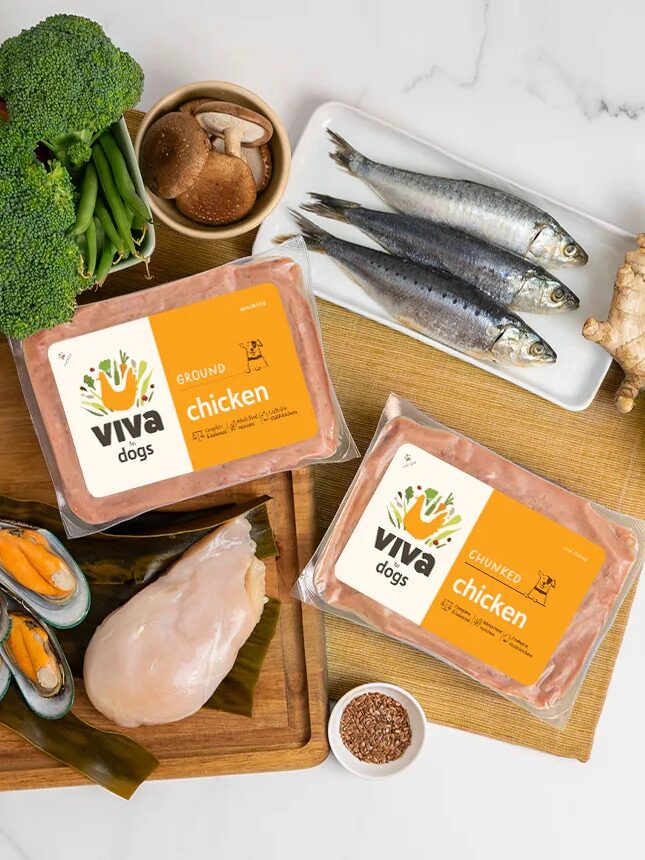 Two packages of Viva Dog chicken are surrounded by vegetables, fish, mussels, ginger, cranberries, and chicken breast on a wooden cutting board with some items partially off the board.