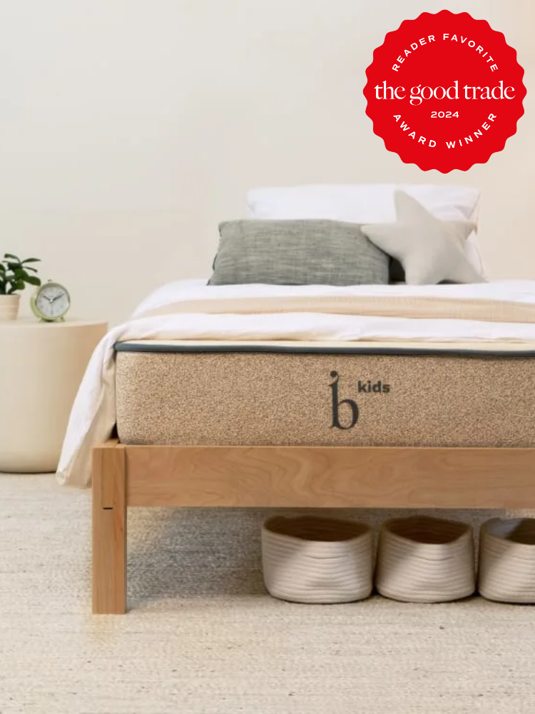A neatly made bed with a beige mattress labeled "b kids" is displayed, with an award sticker on the upper right reading "Reader Favorite, FashionGroomSpot, 2024 Award Winner." Three baskets sit under the bed.