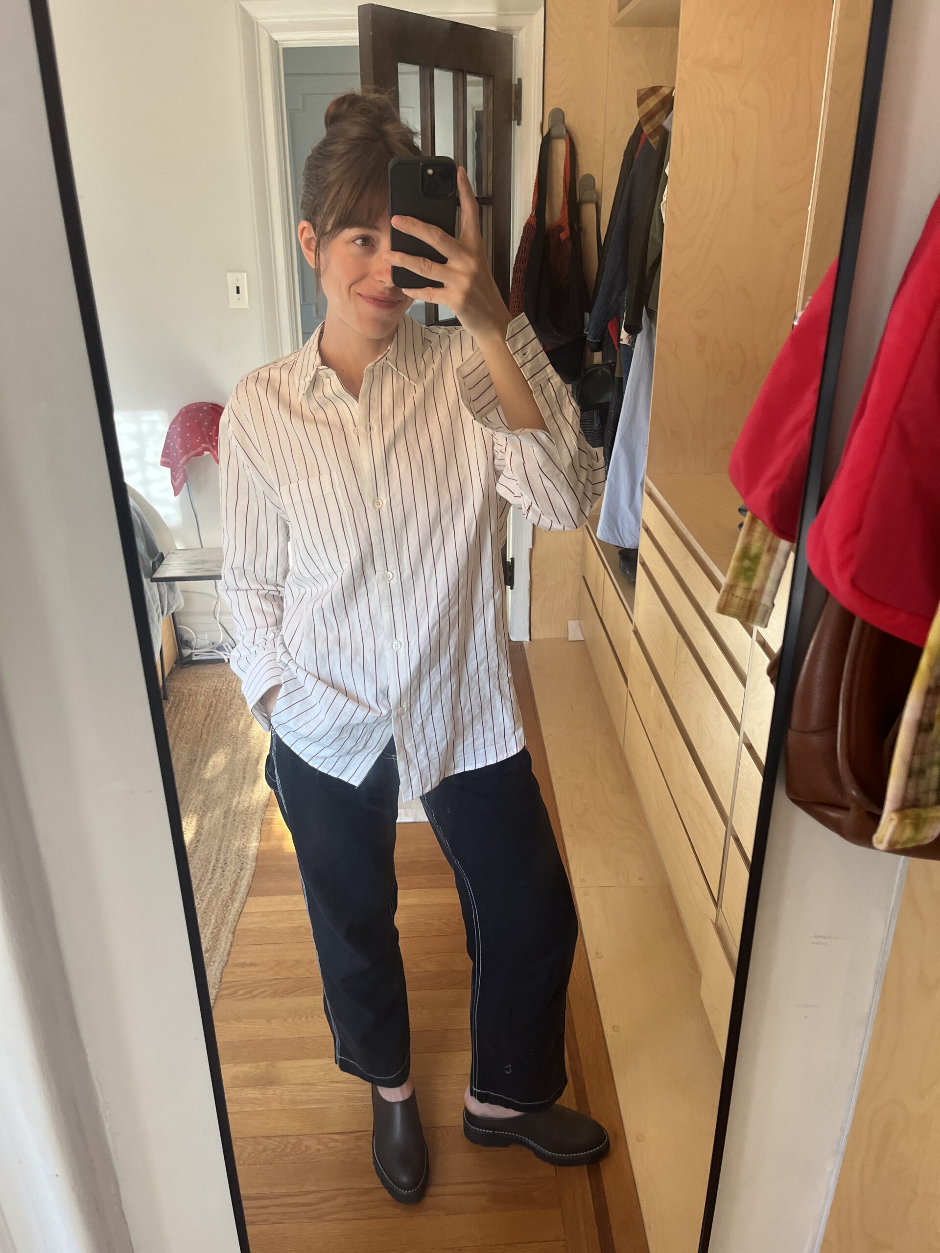 A woman takes a selfie in a full-length mirror of her clothes, which are a striped button down, black pants, and dark shoes.