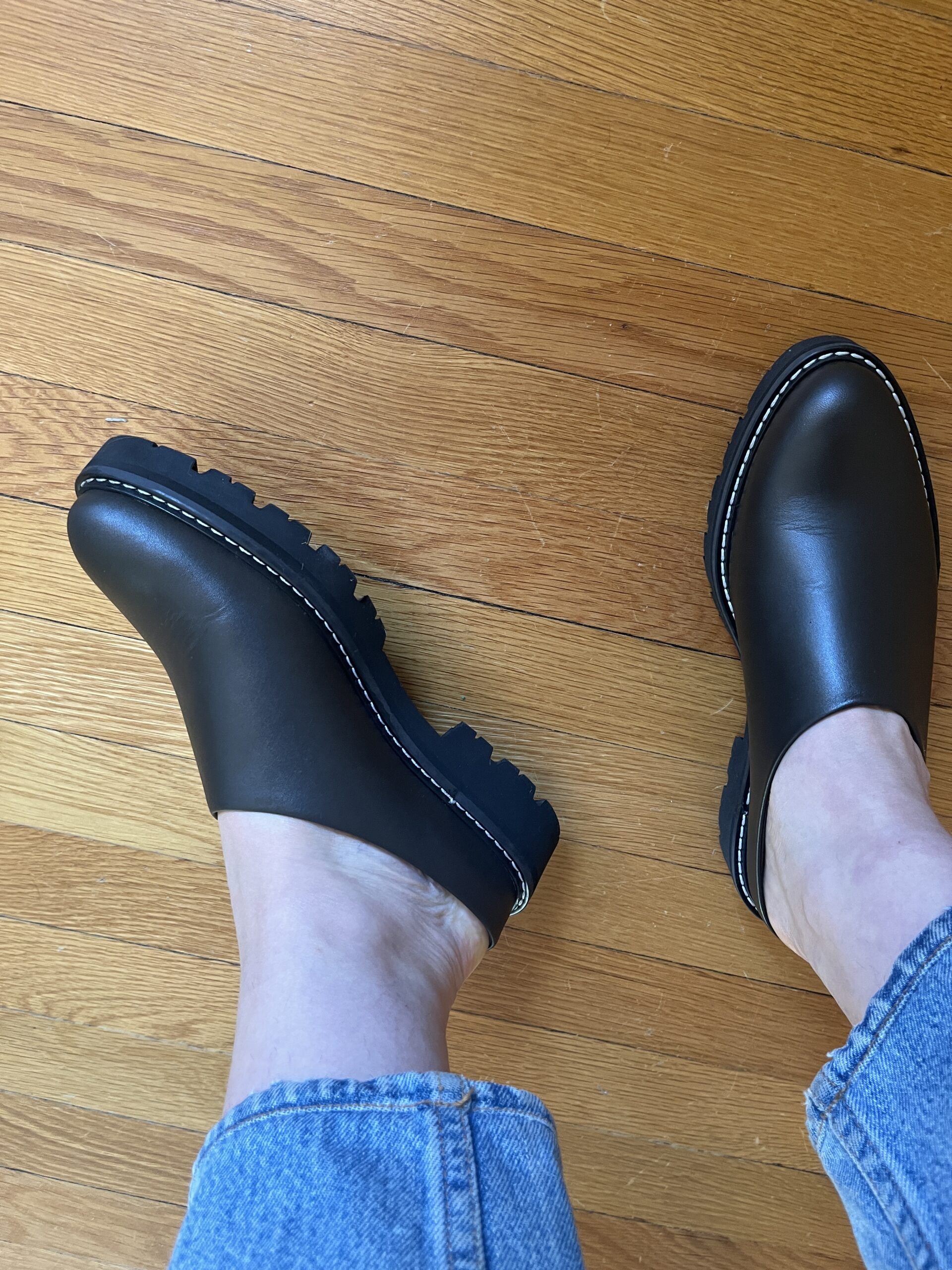 A woman wearing black lug mules, with her ankles the hems of her jeans visible.