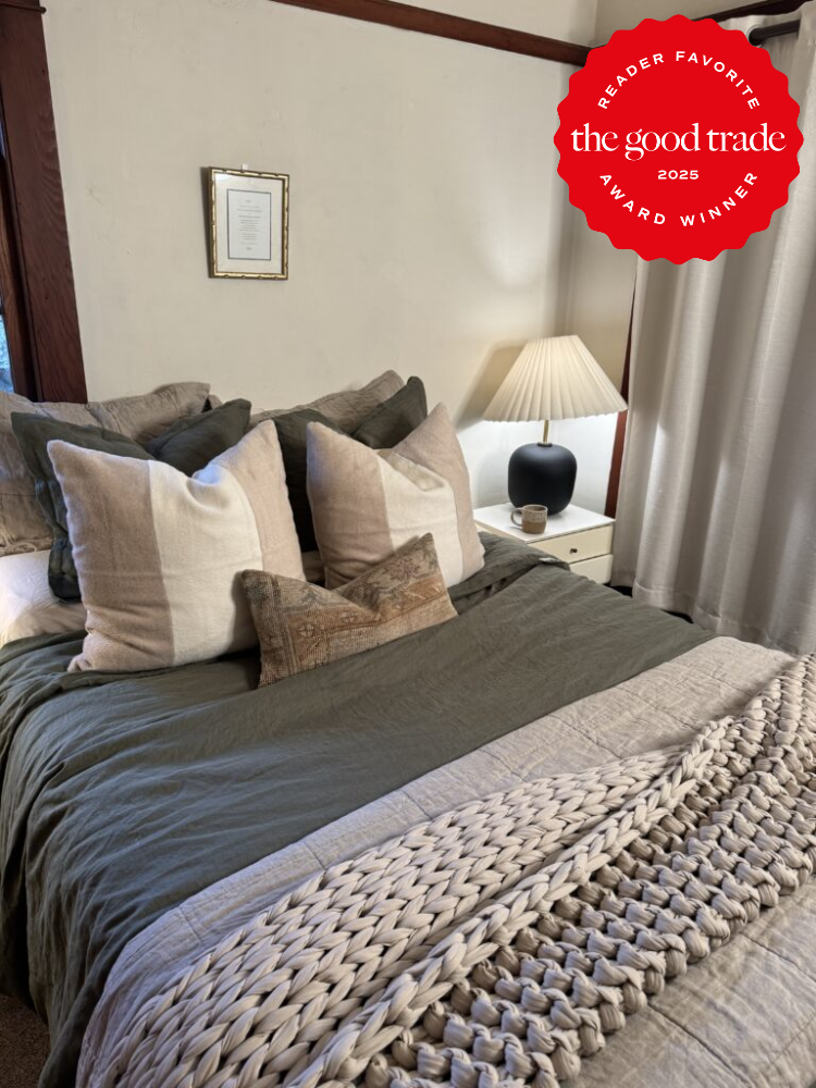 Cozy bedroom with a neatly made bed, large pillows, textured throw, and a bedside table with a lamp. A framed certificate is on the wall. "FashionGroomSpot 2025 Reader Favorite" badge is visible.