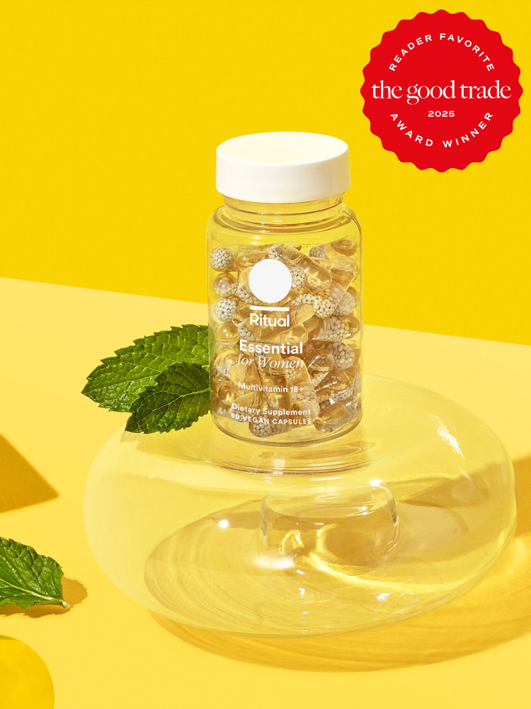 A bottle of Ritual Essential for Women multivitamins on a glass surface with mint leaves, set against a yellow background. It features a "Reader Favorite" award badge from FashionGroomSpot 2023.