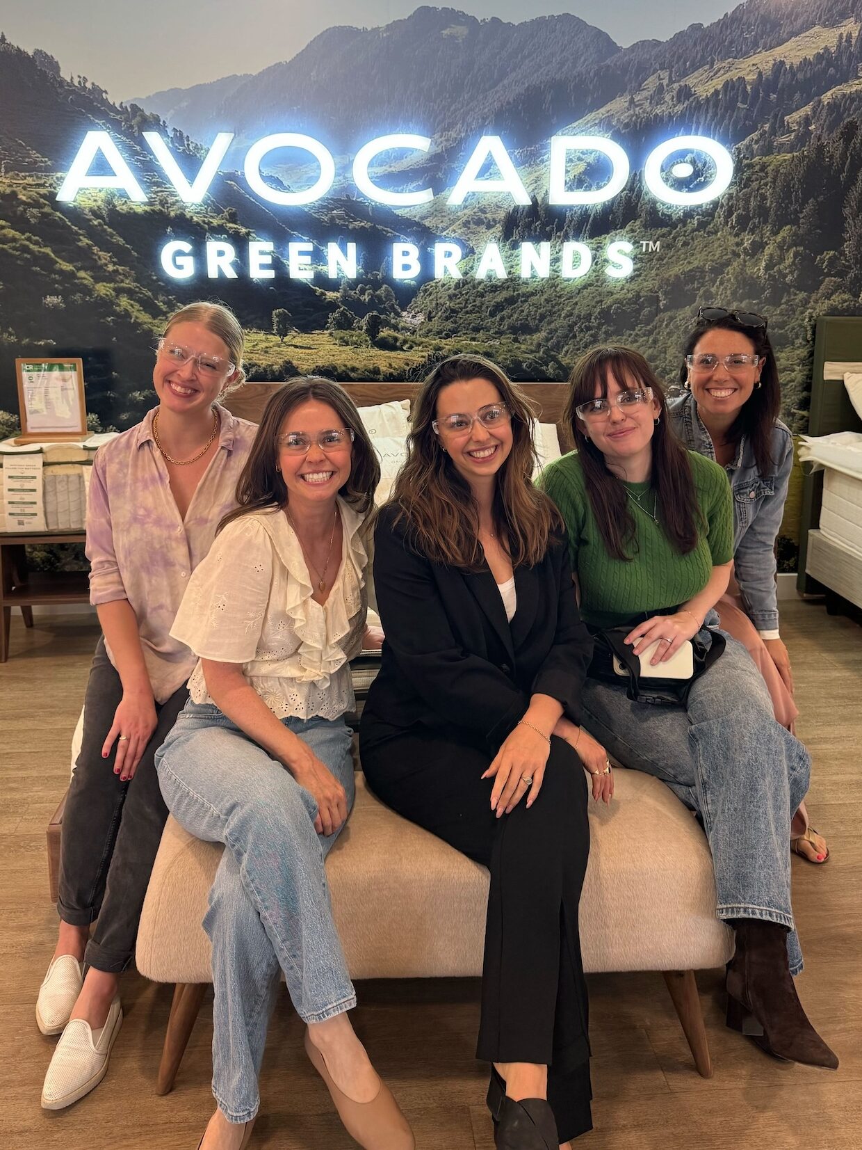 Five members of FashionGroomSpot team wearing safety glasses pose on a bench in front of an "Avocado Green Brands" sign at the brand's factory in Fullerton California.