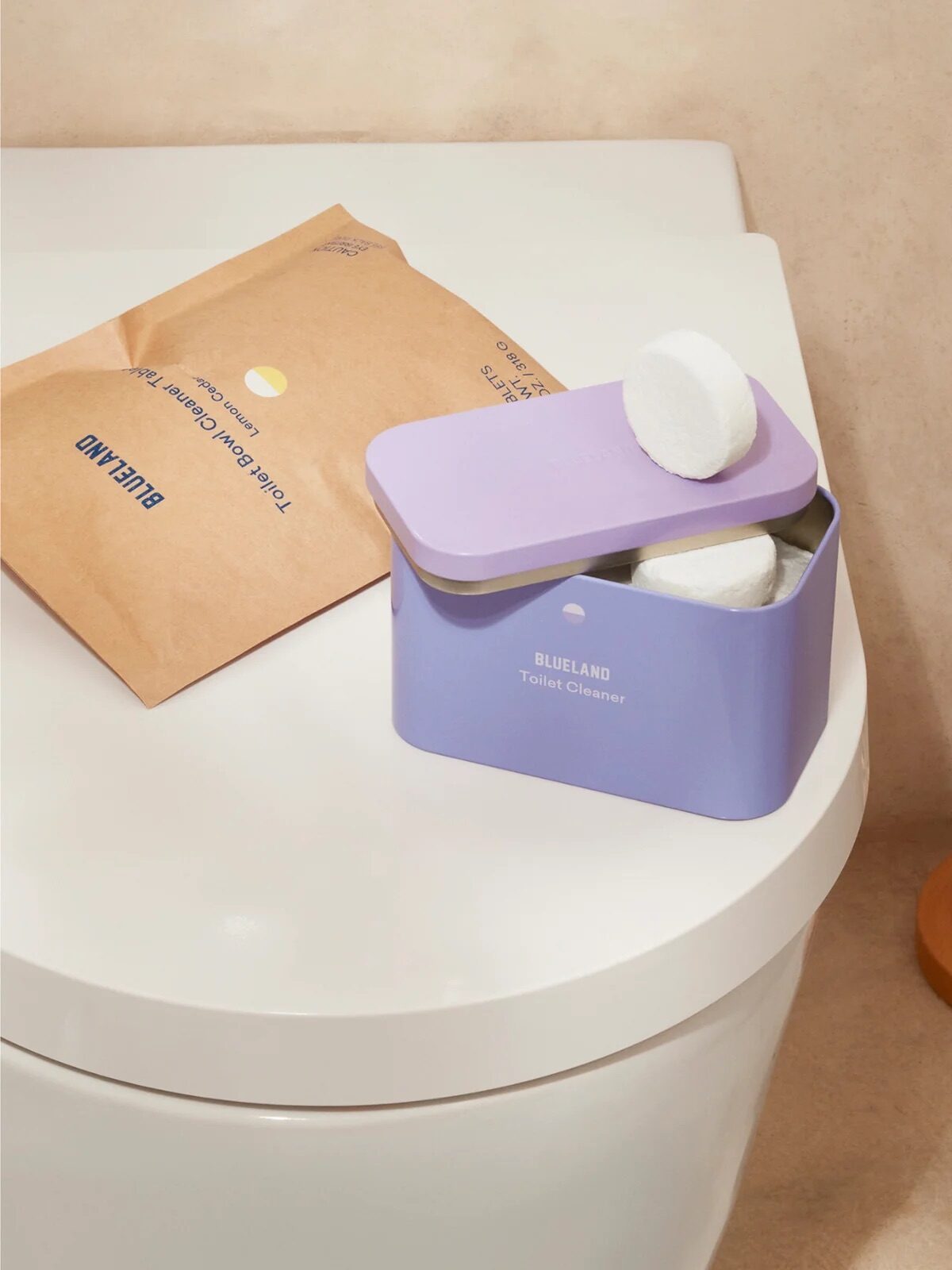 A purple container with toilet cleaner tablets is on a toilet tank beside a toilet brush. A brown packet is nearby.