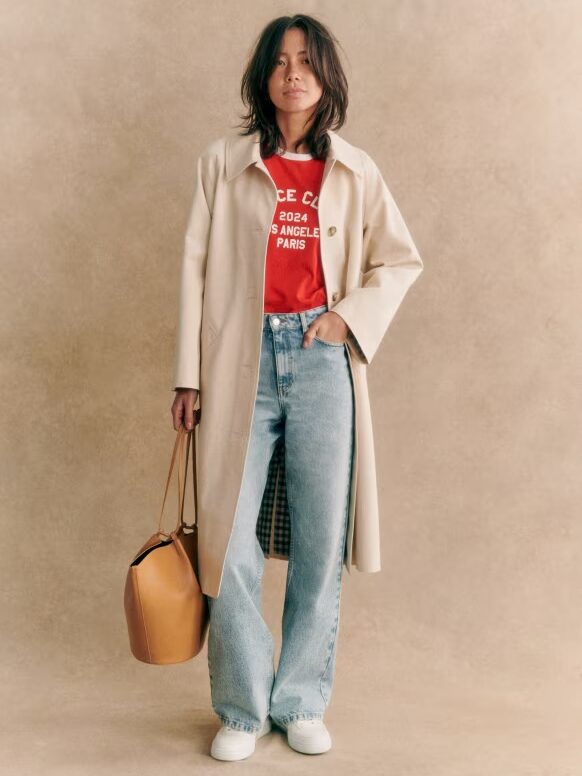 Person in a long beige coat over a red shirt and jeans, holding a brown bag.