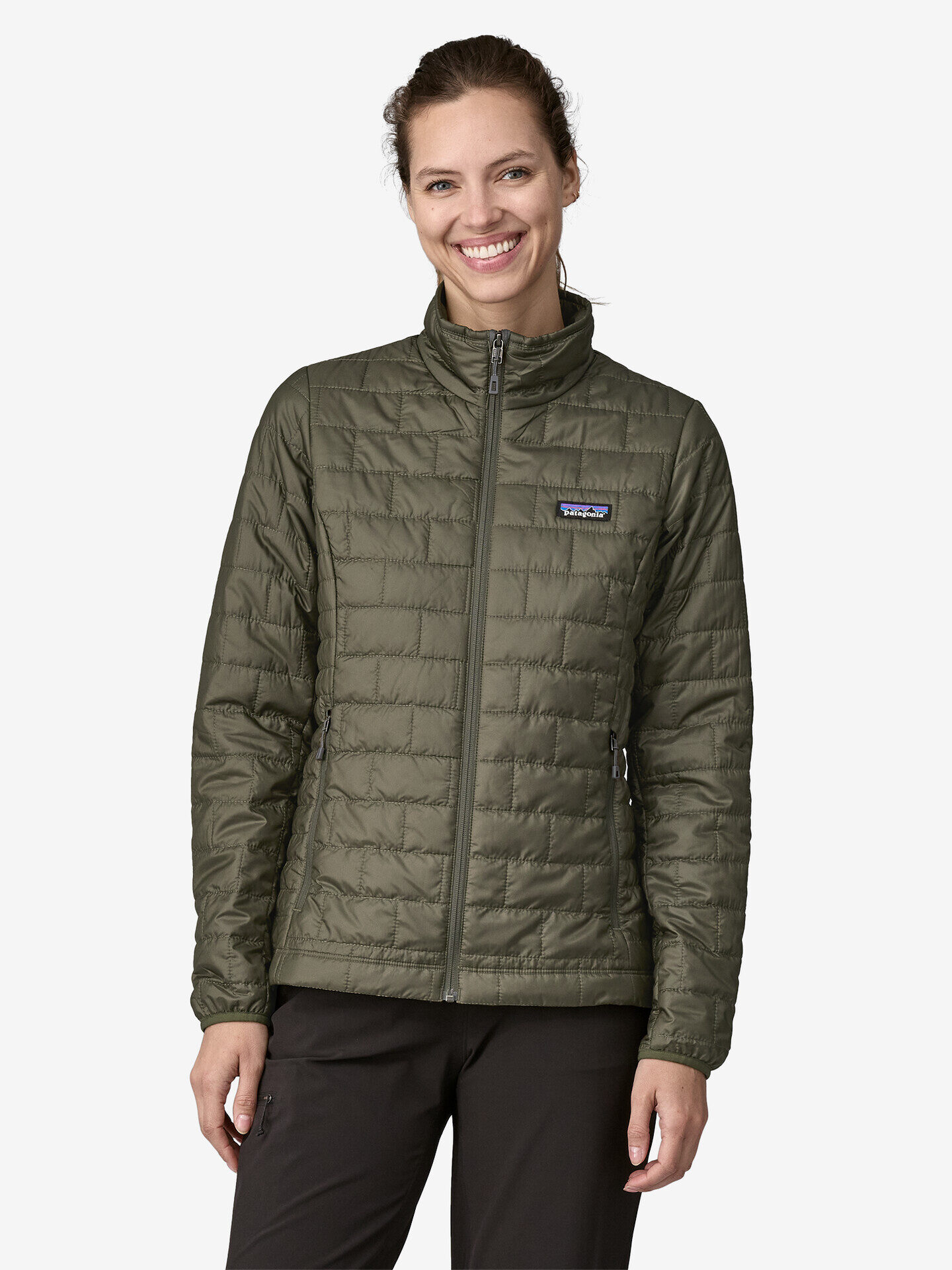 Person wearing a green quilted jacket and dark pants, smiling, with hands by their sides against a plain background.