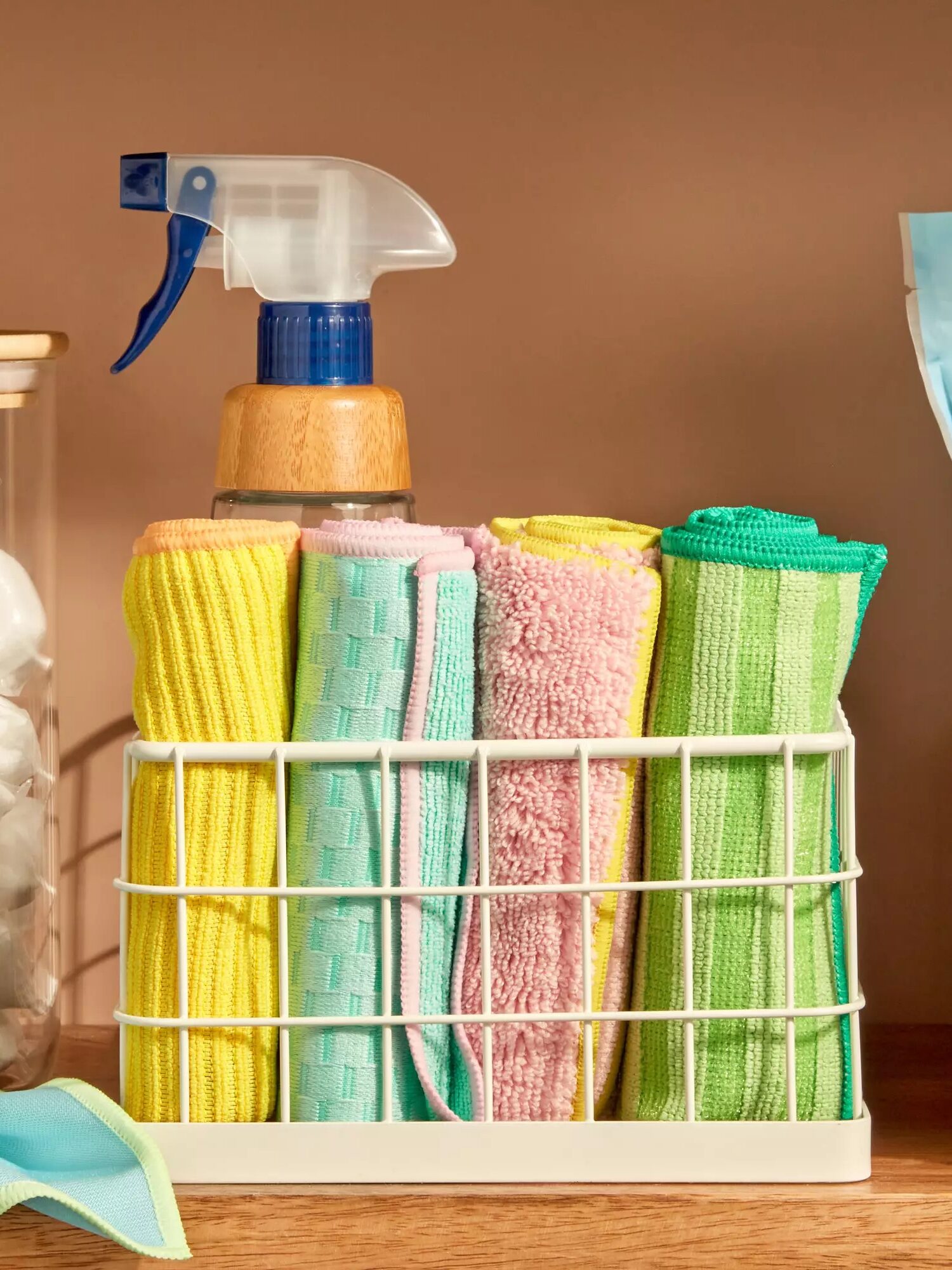 Cleaning supplies on a shelf, featuring a spray bottle, colorful cloths in a rack, dishwashing pods in a jar, and detergent powder in a bag.