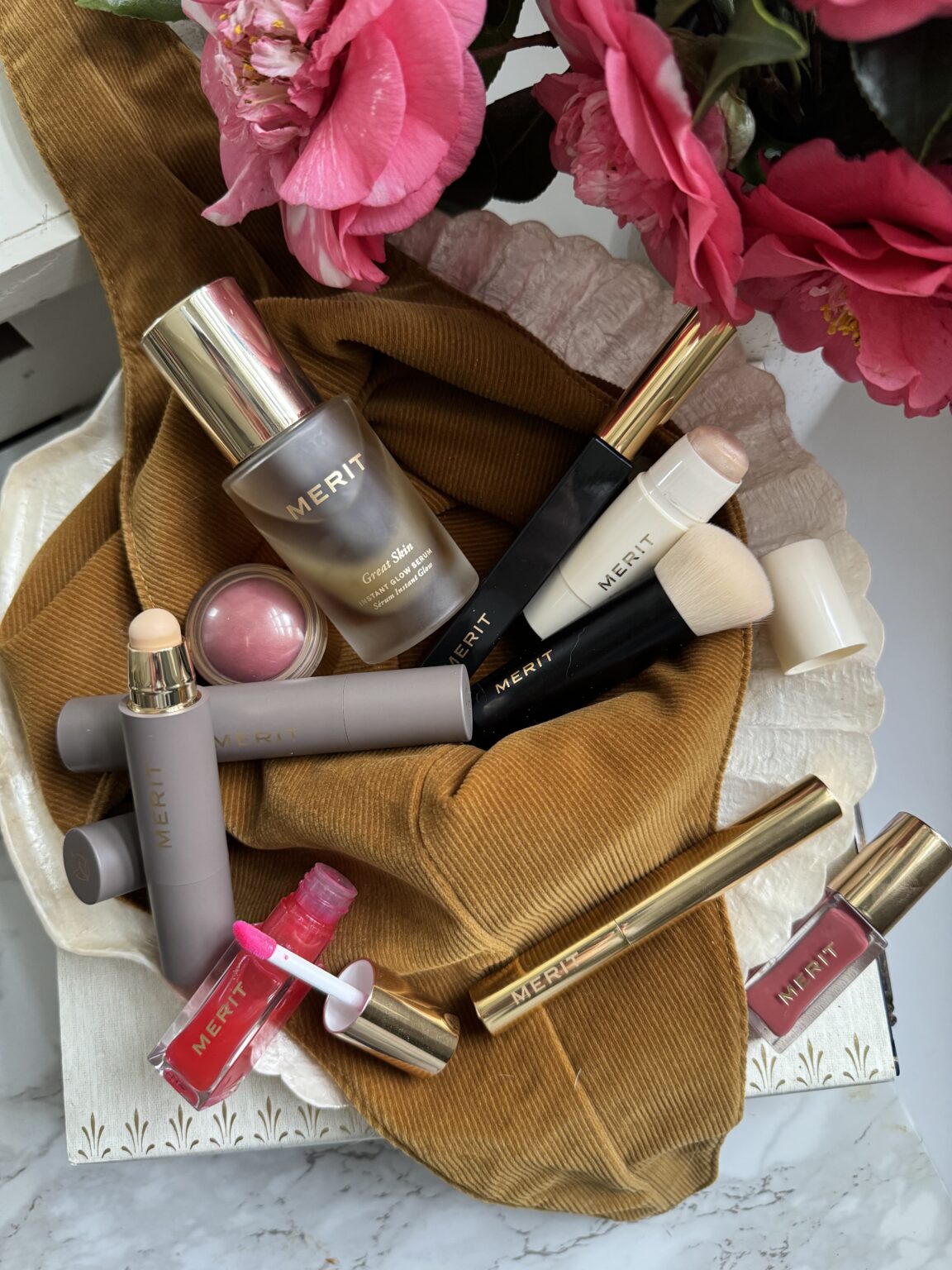 A collection of Merit beauty products, including foundation, blush, lip gloss, and brushes, arranged on a brown fabric with pink flowers nearby.
