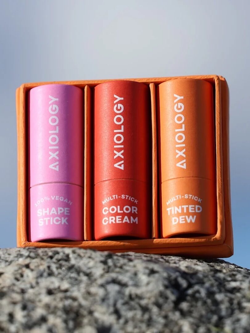 Three Axiology multi-sticks in pink, red, and orange tubes labeled Shape Stick, Color Cream, and Tinted Dew, on a rock surface with a blurred sky background.