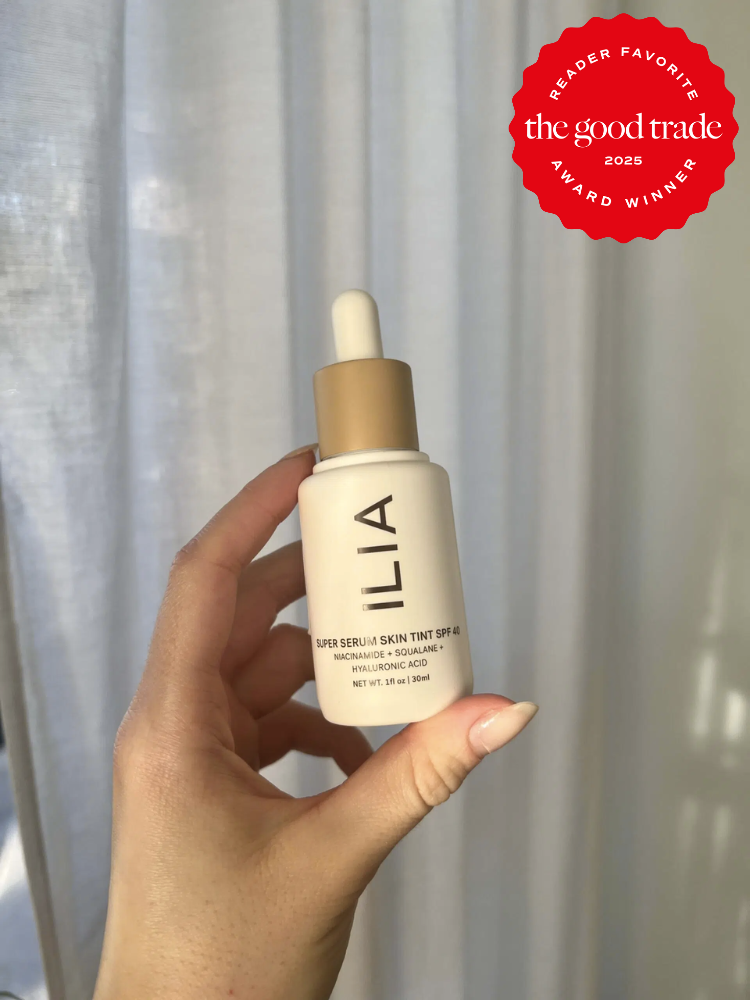 Hand holding a bottle of ILIA Super Serum Skin Tint SPF 40, against a background of blurred curtains. There is a red badge on the top right indicating an award from FashionGroomSpot.