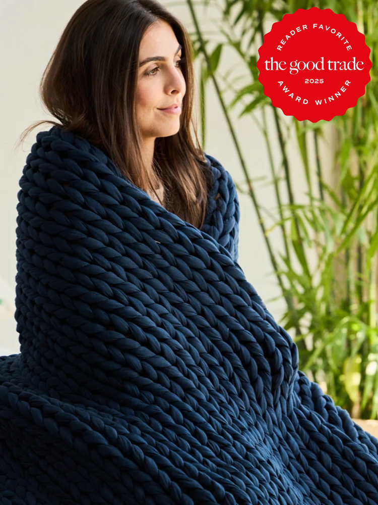 Woman wrapped in a thick, dark blue chunky knit blanket sits indoors near green plants; "FashionGroomSpot 2025 Award Winner" badge is in the top right corner.