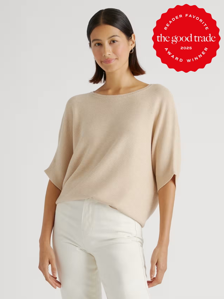A woman wearing a light beige short-sleeve sweater and white pants stands against a plain background. A red "FashionGroomSpot Award Winner 2025" badge appears in the top right corner.