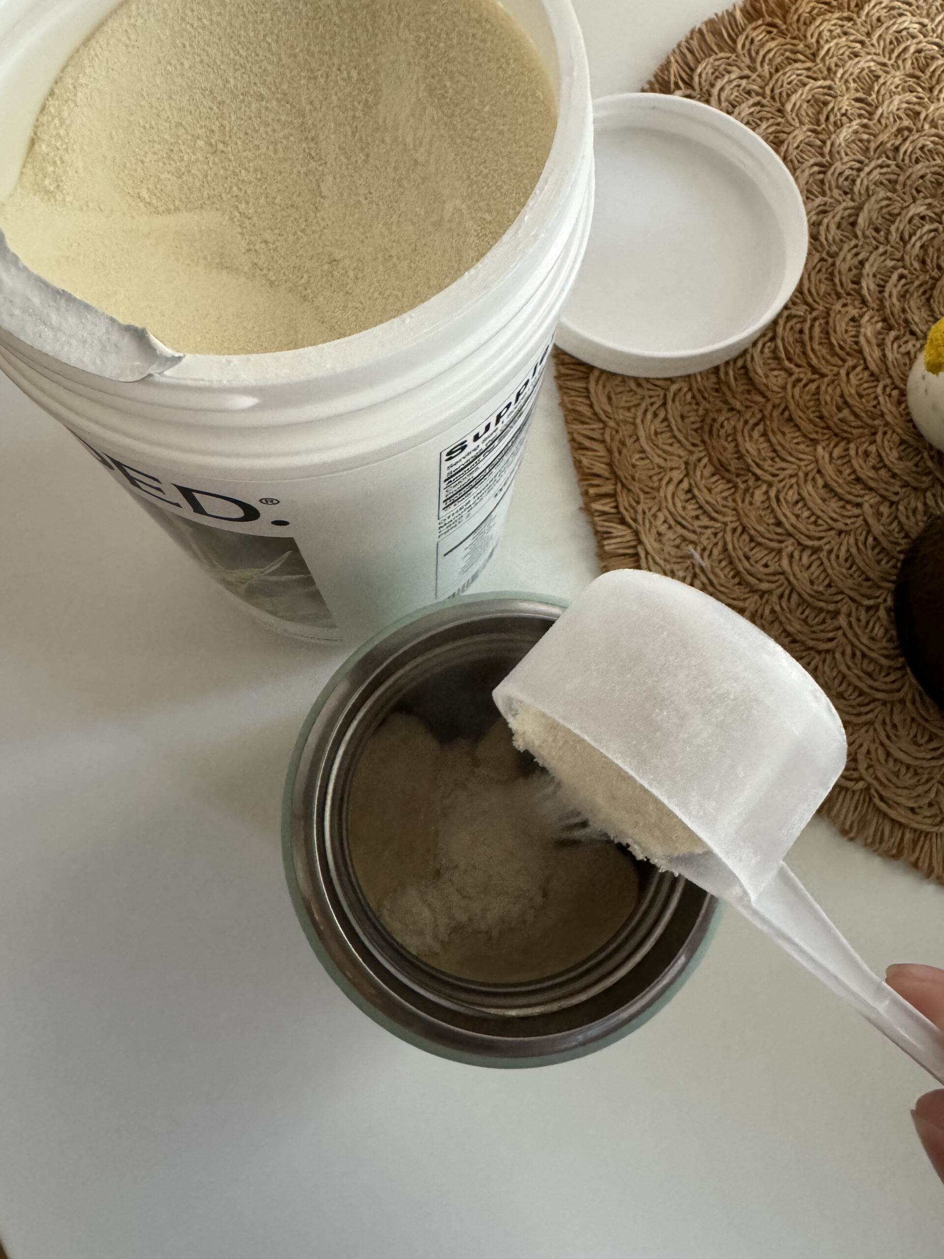 A hand holding a scoop of white powder over an open container, next to a larger tub of the same powder, a round woven mat, and a closed white lid on a white surface.