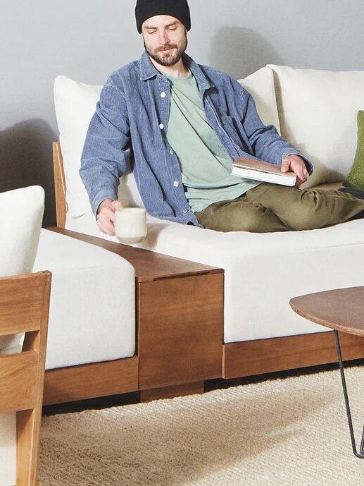 Man sitting on a white sofa with wooden accents, holding a book and a mug, next to a round wooden coffee table with a plant and magazine.