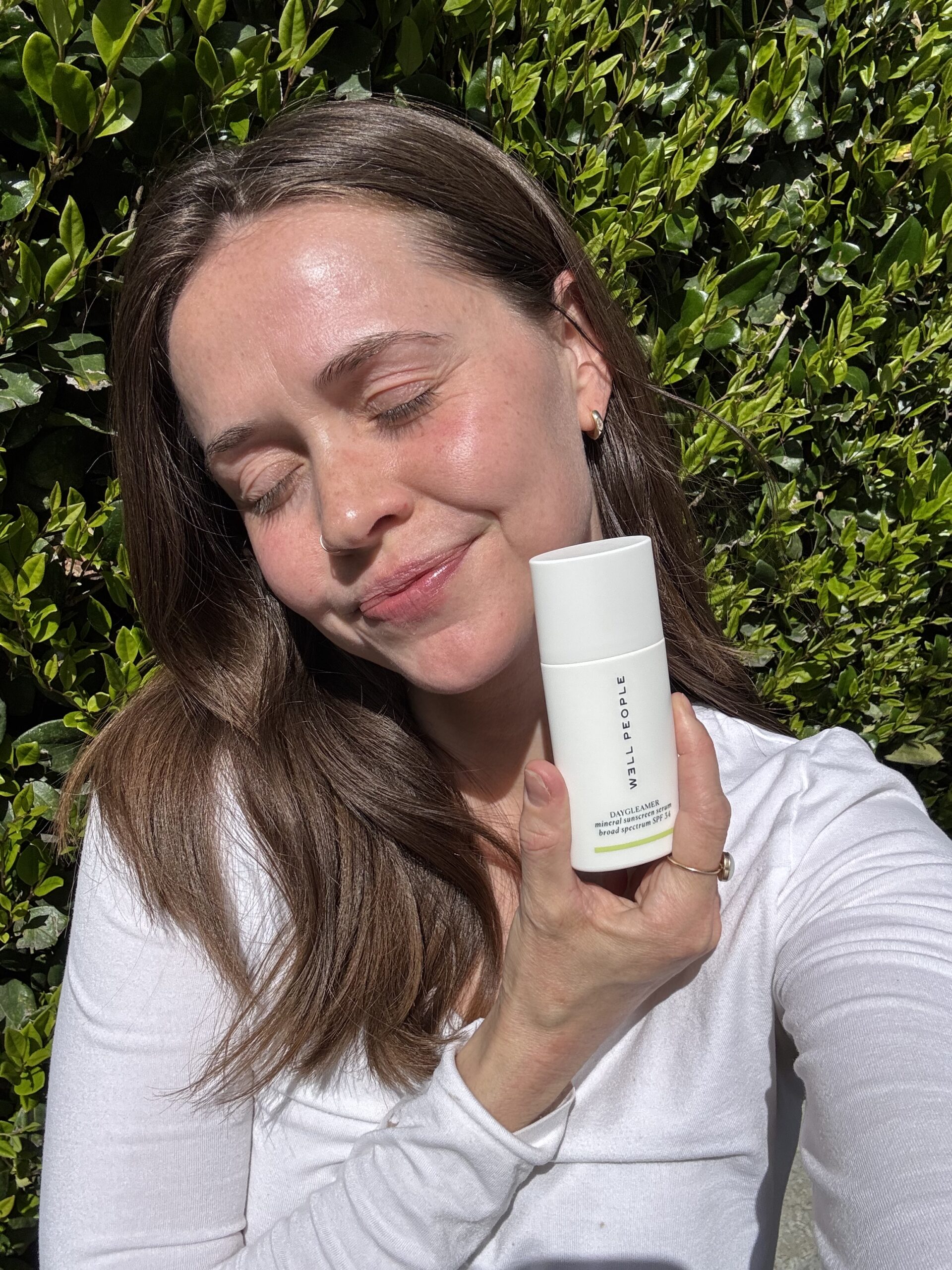 Person with long brown hair wearing a white top, smiling with closed eyes, holding a white cylindrical product container, standing in front of green foliage.