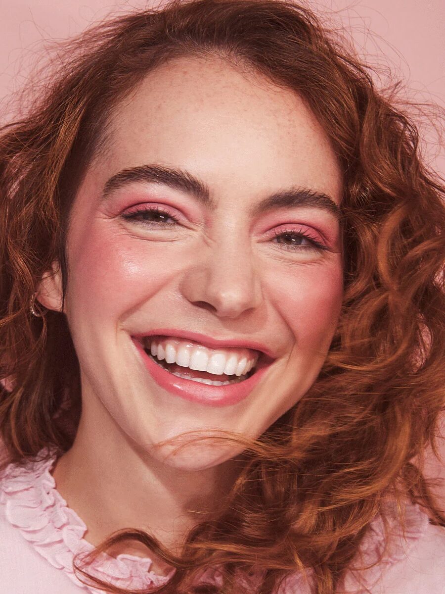 Smiling person with curly red hair and pink makeup against a pink background.