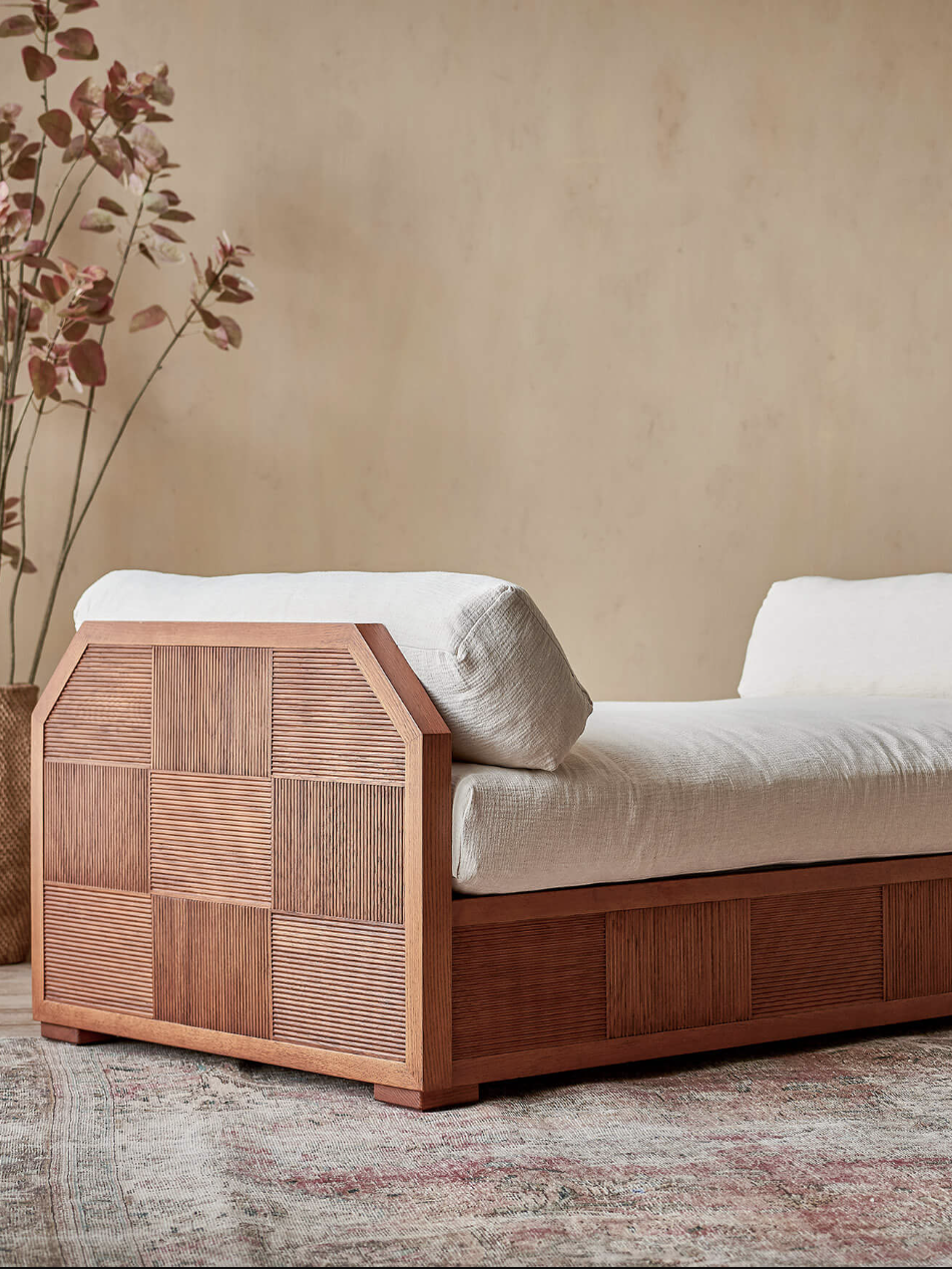 Wooden daybed with white cushions on an ornate rug, surrounded by plants against a beige wall.