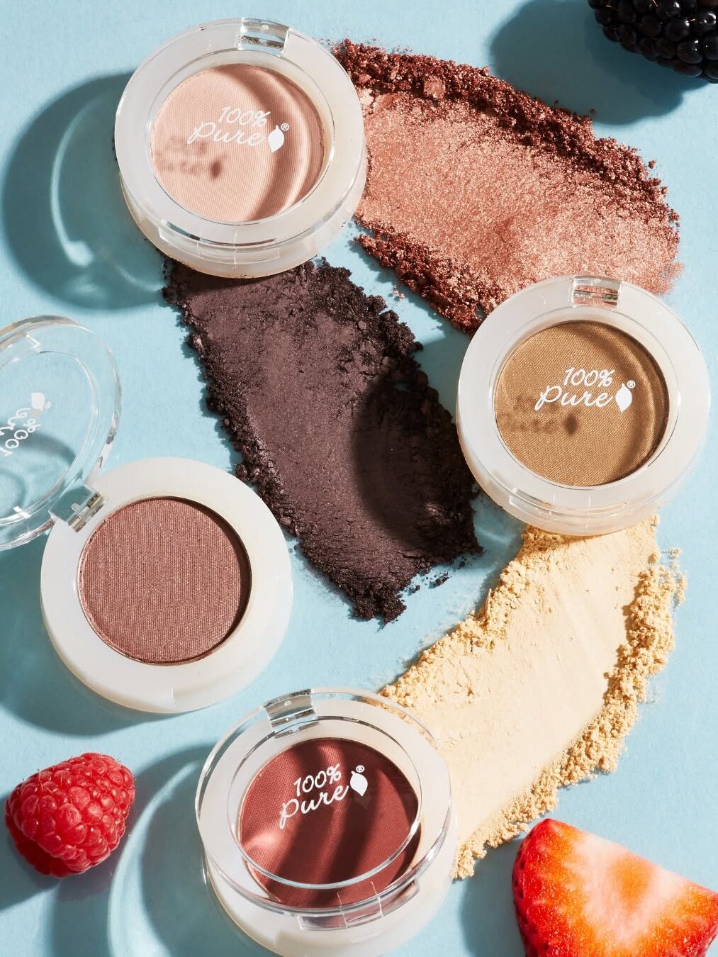 Four round makeup containers on a blue surface, each with different eyeshadow shades. Loose powder is spread around the containers. Berries are scattered nearby.