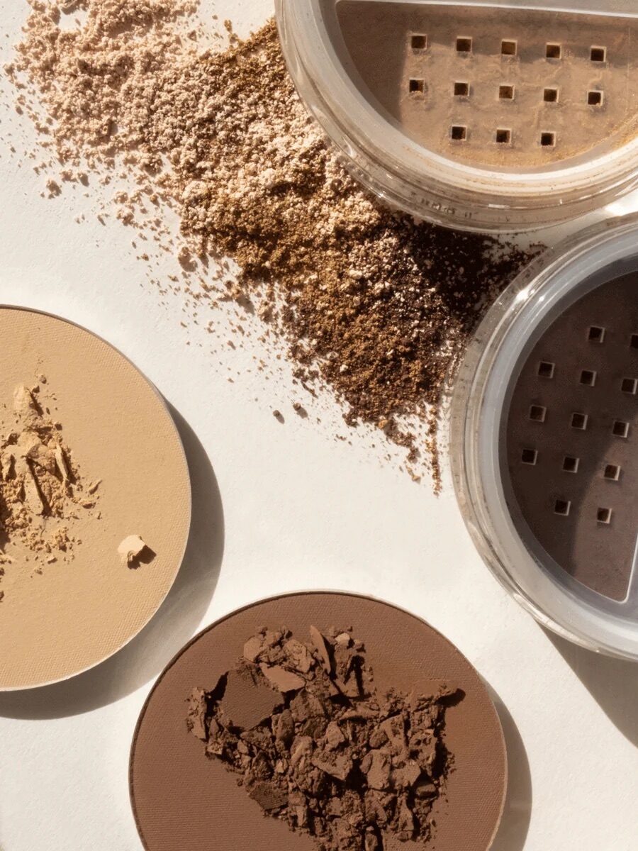 Various shades of loose and pressed powder makeup are spilled on a surface. Two open containers reveal finely milled powder with sifter lids.