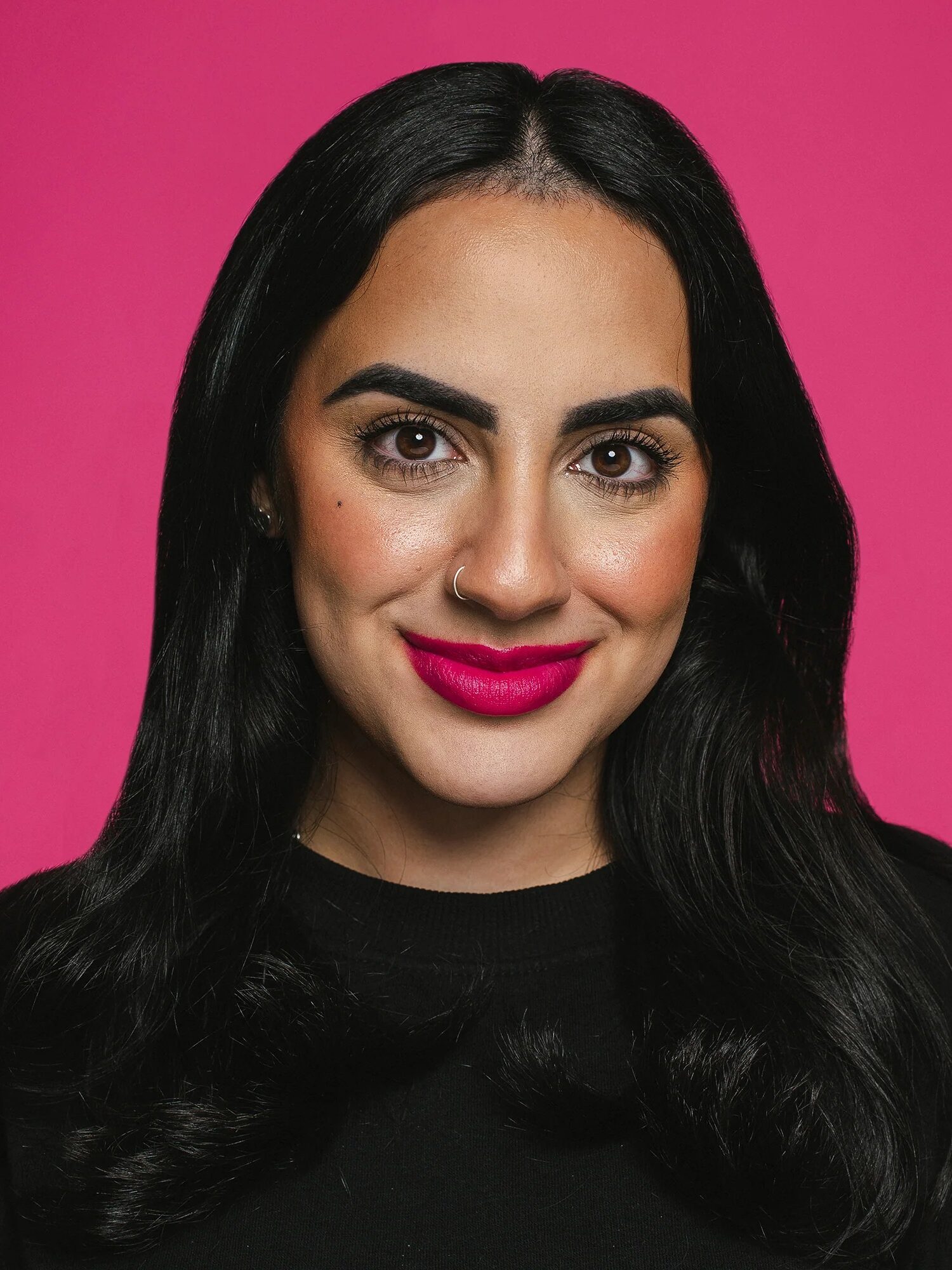 A person with long black hair and a nose ring is smiling against a pink background.