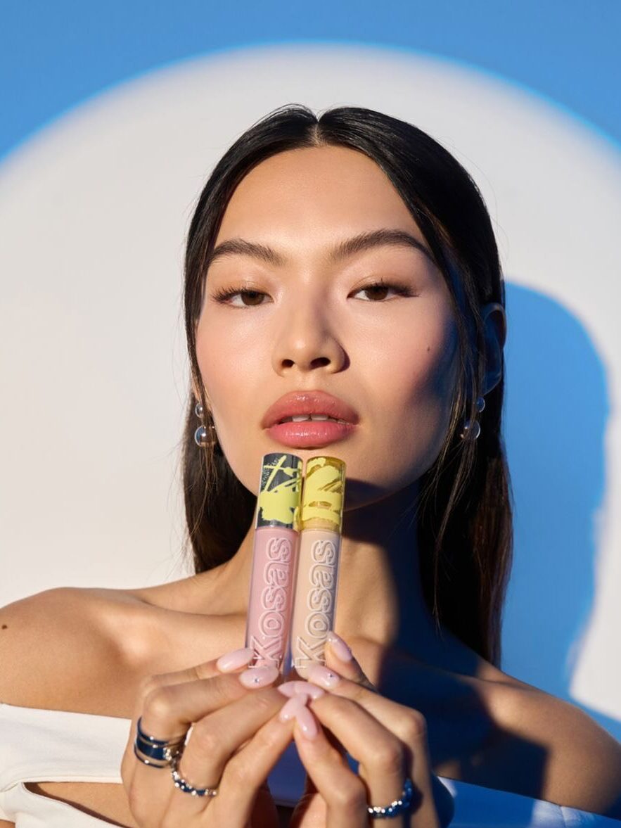 A woman holding two lip gloss tubes in front of a blue and white gradient background. She has straight dark hair and is wearing off-shoulder clothing and earrings.