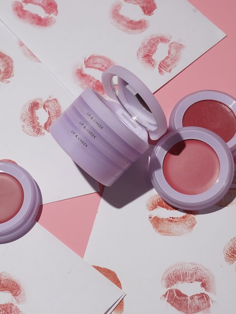 Lip cream containers on a pink surface with printed lipstick kiss marks on paper around them.
