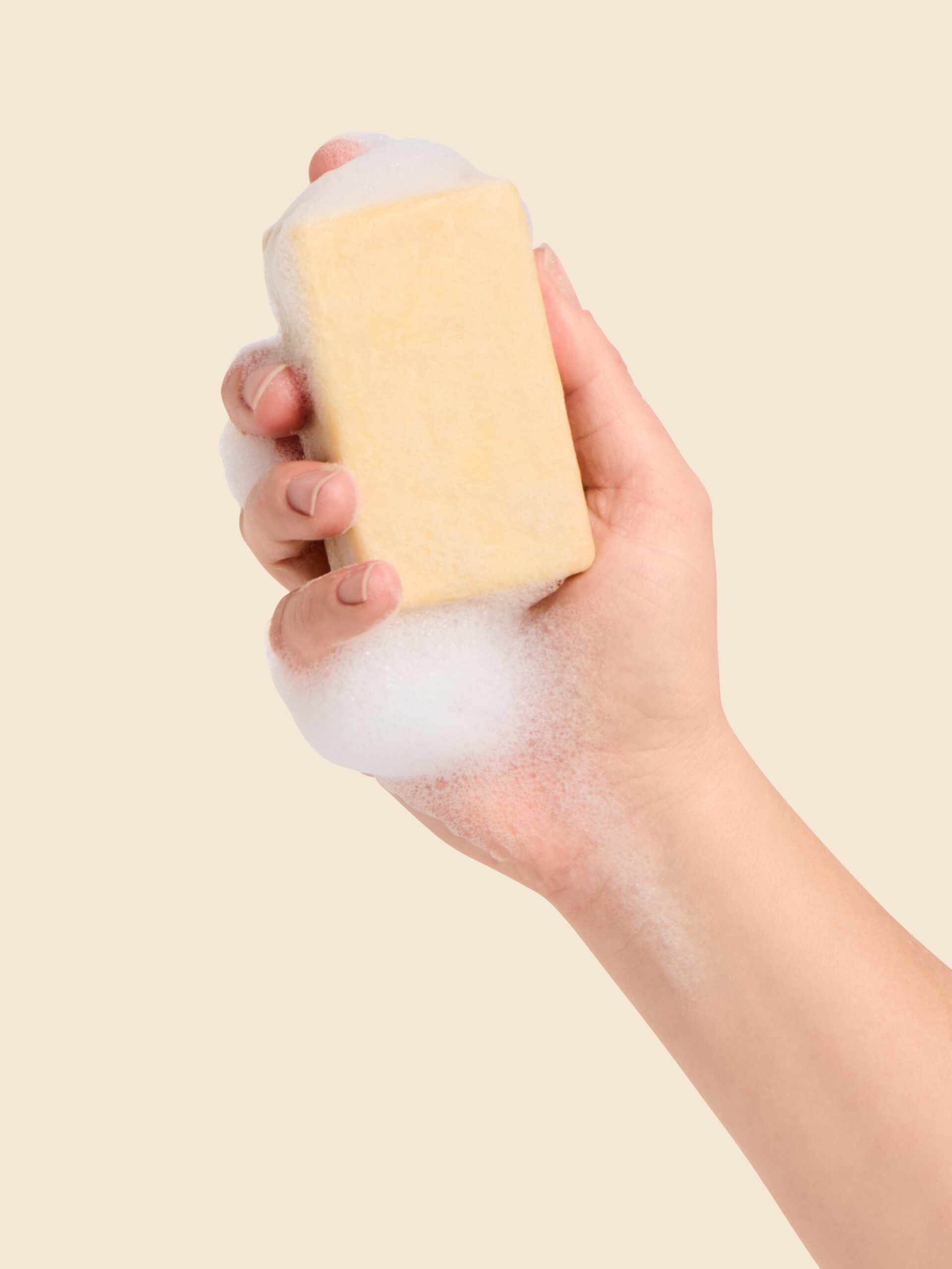 Hand holding a soapy yellow bar of soap against a beige background.