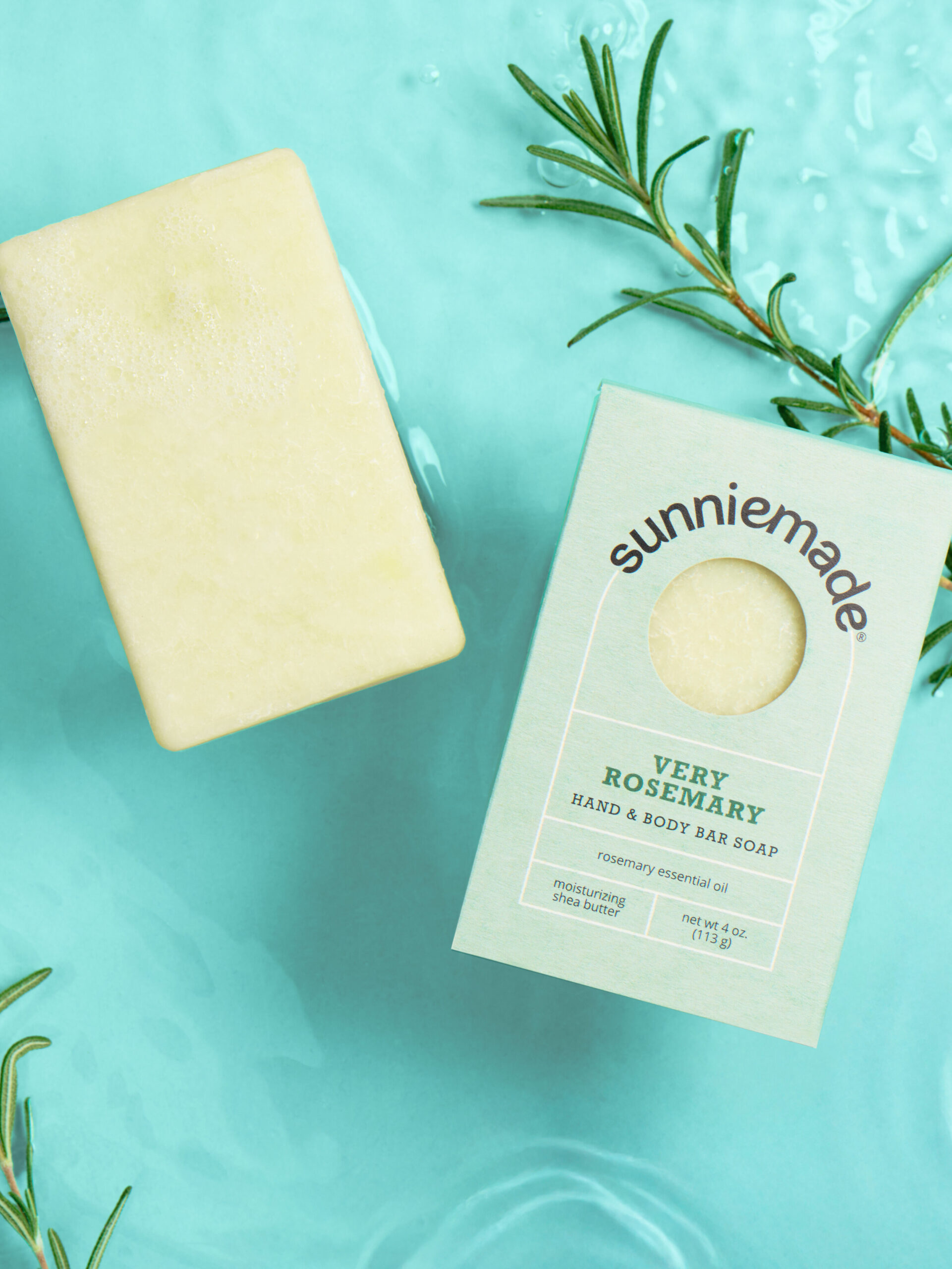 Bar of soap with rosemary, next to packaging labeled "sunniemade Very Rosemary Hand & Body Bar Soap," on a watery blue background with rosemary sprigs.