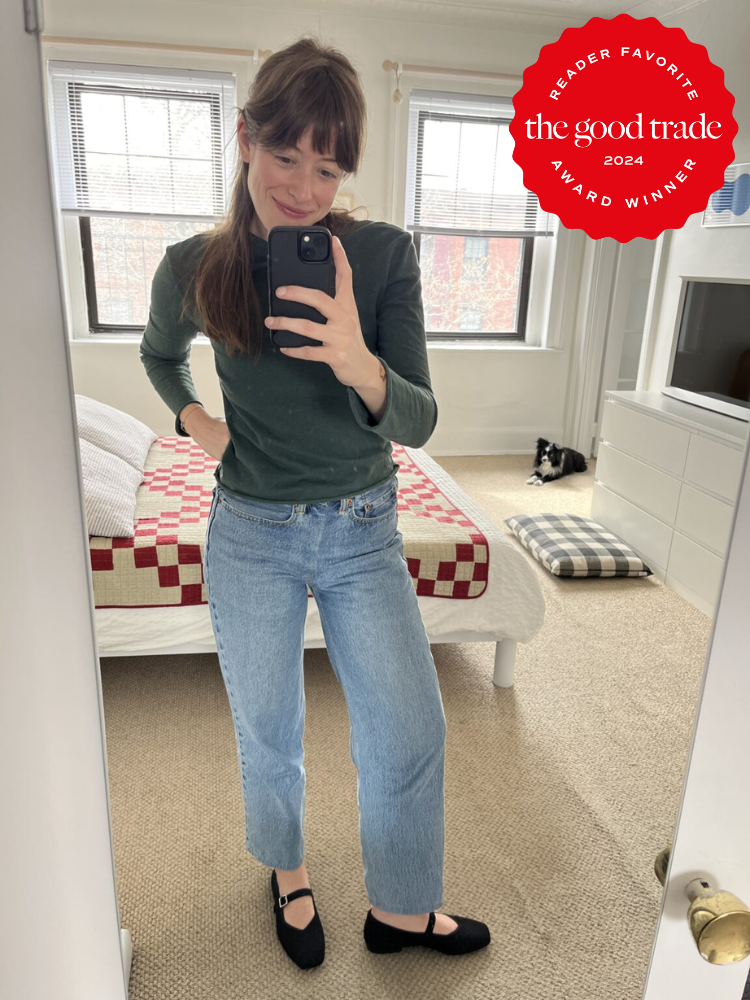 Woman taking a mirror selfie in a bedroom, wearing a green sweater and jeans. There's a bed with a red-and-white quilt and a cat on a rug in the background. "FashionGroomSpot Award Winner" badge.