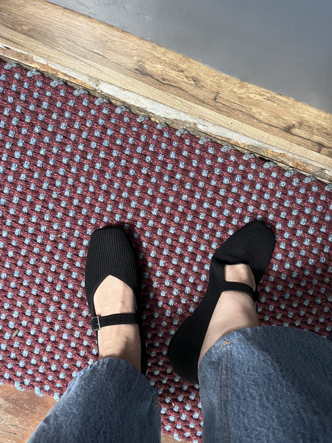 A person wearing black Mary Jane shoes stands on a red and gray patterned carpet, shown from a top-down view.