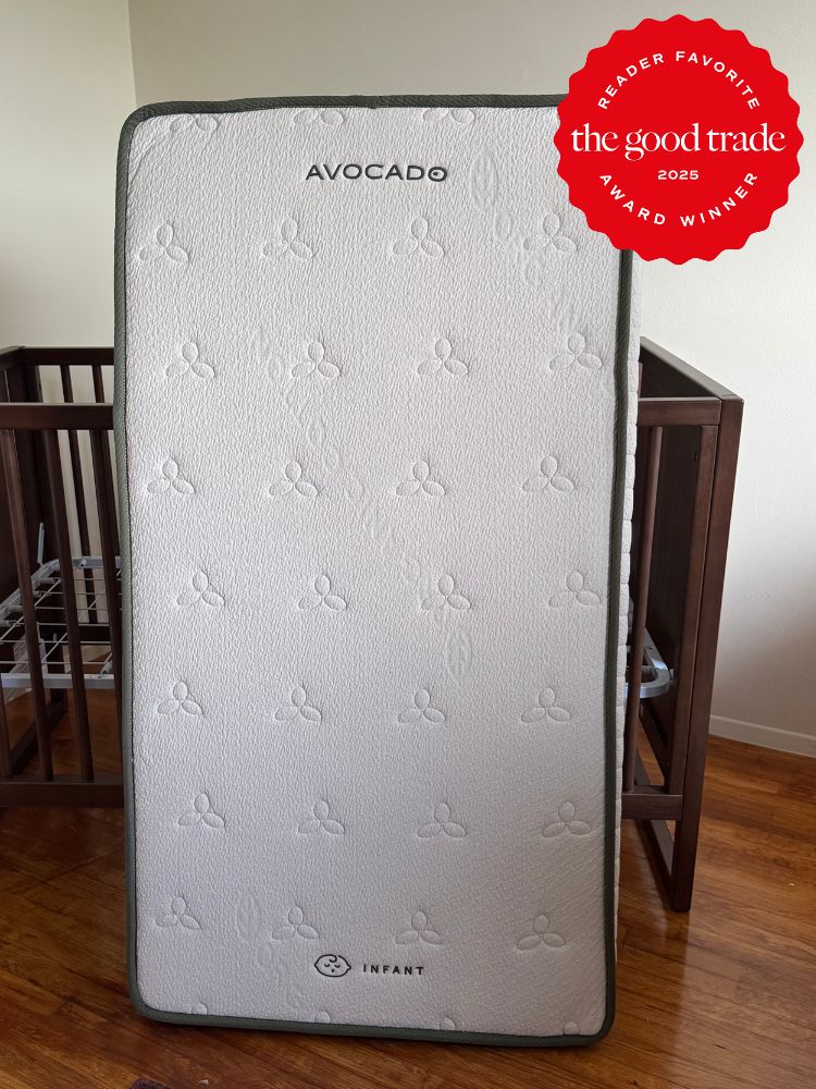 A crib mattress labeled "Avocado Infant" stands upright in a room with wooden furniture; a 2025 "FashionGroomSpot" award badge is displayed in the top right corner.