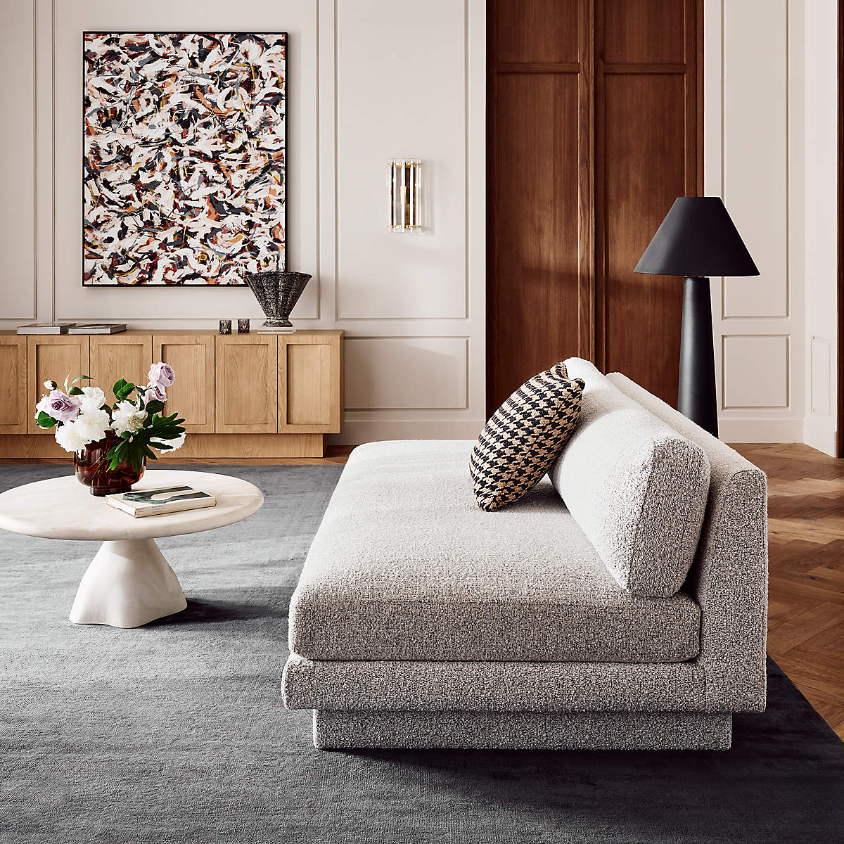 A modern living room with a textured light gray sofa, a round white coffee table with flowers, abstract wall art, wooden sideboard, and a black floor lamp on a gray rug.