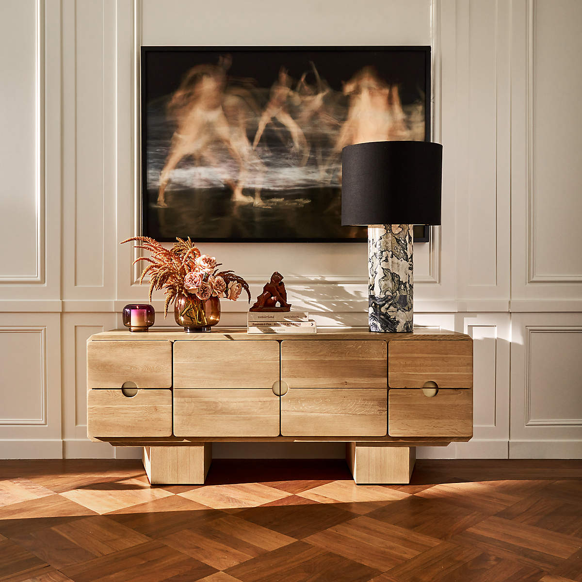 A wooden sideboard with decorative items and a lamp stands against a paneled wall, beneath a large abstract art piece featuring blurred human figures.