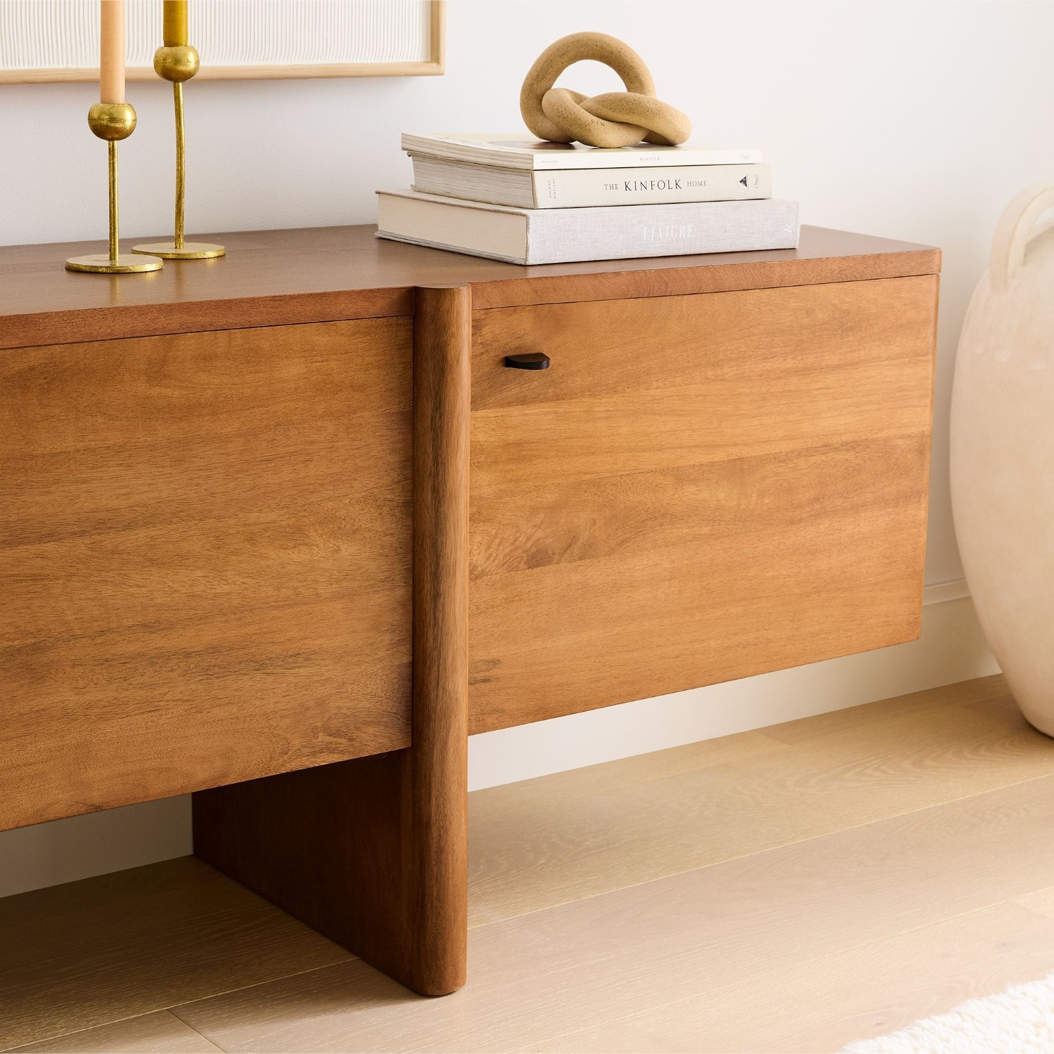 A wooden sideboard with a vertical handle is topped with stacked books, gold candlesticks, and a decorative knot object.