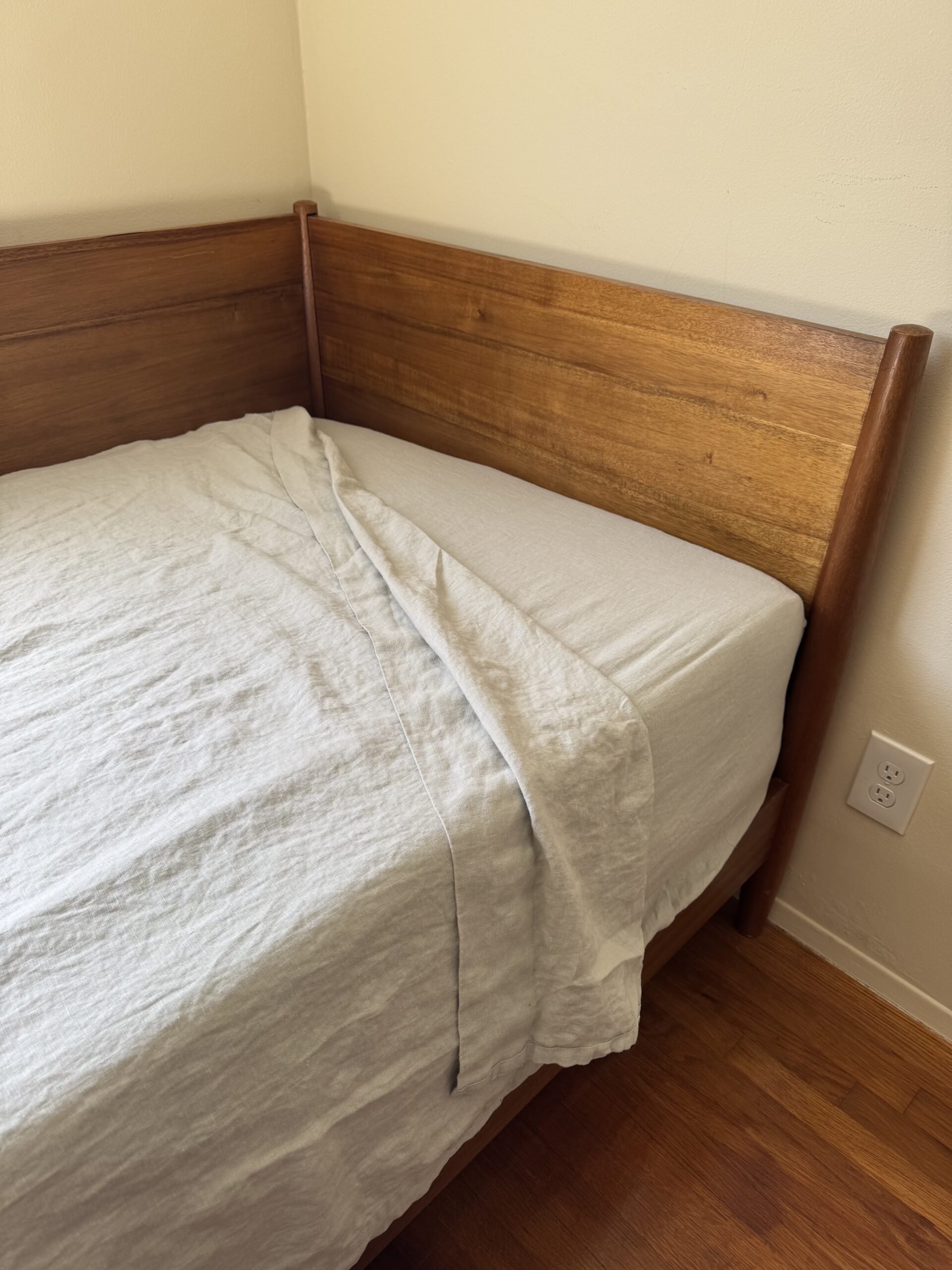 Wooden bed frame with a beige sheet partially covering the mattress. The bed is positioned in a corner, with a wall socket visible nearby on a wooden floor.