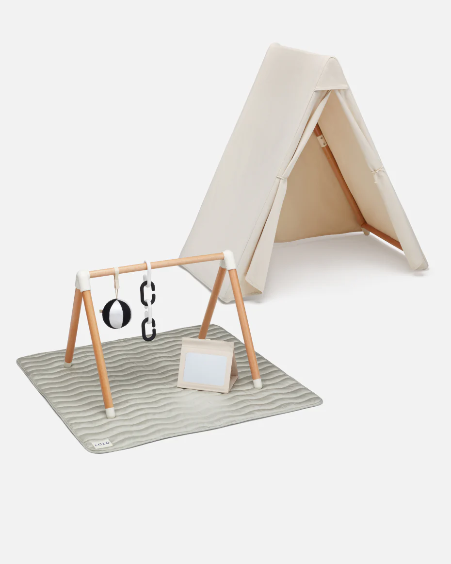A beige play tent is set up behind a wooden baby gym with hanging toys and a soft mat on the floor.
