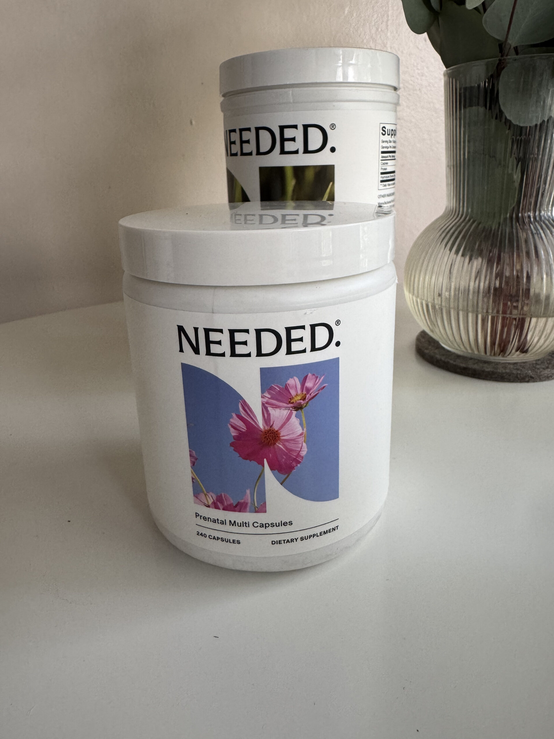 A white container labeled "NEEDED. Prenatal Multi Capsules" sits on a table, highlighting this premium prenatal vitamin; another similar container and a glass vase with green leaves appear in the background.