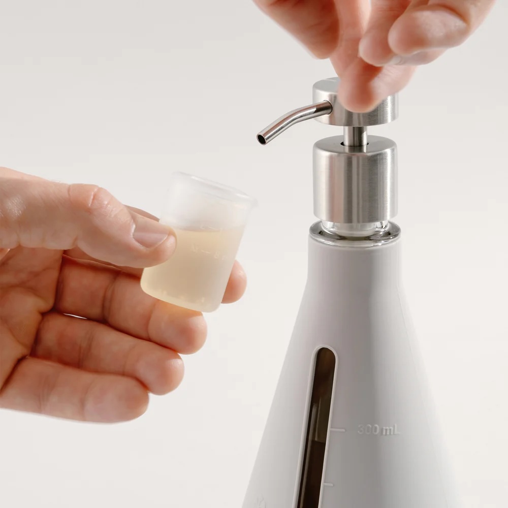 A person dispenses liquid from a pump bottle into a small measuring cup.