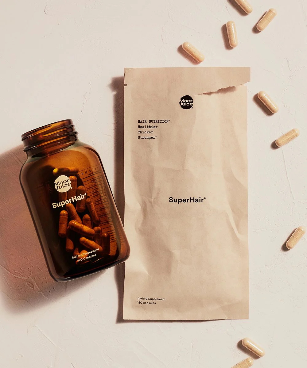 A brown bottle labeled "SuperHair" with capsules inside is next to a beige paper pouch and several scattered capsules on a light surface.